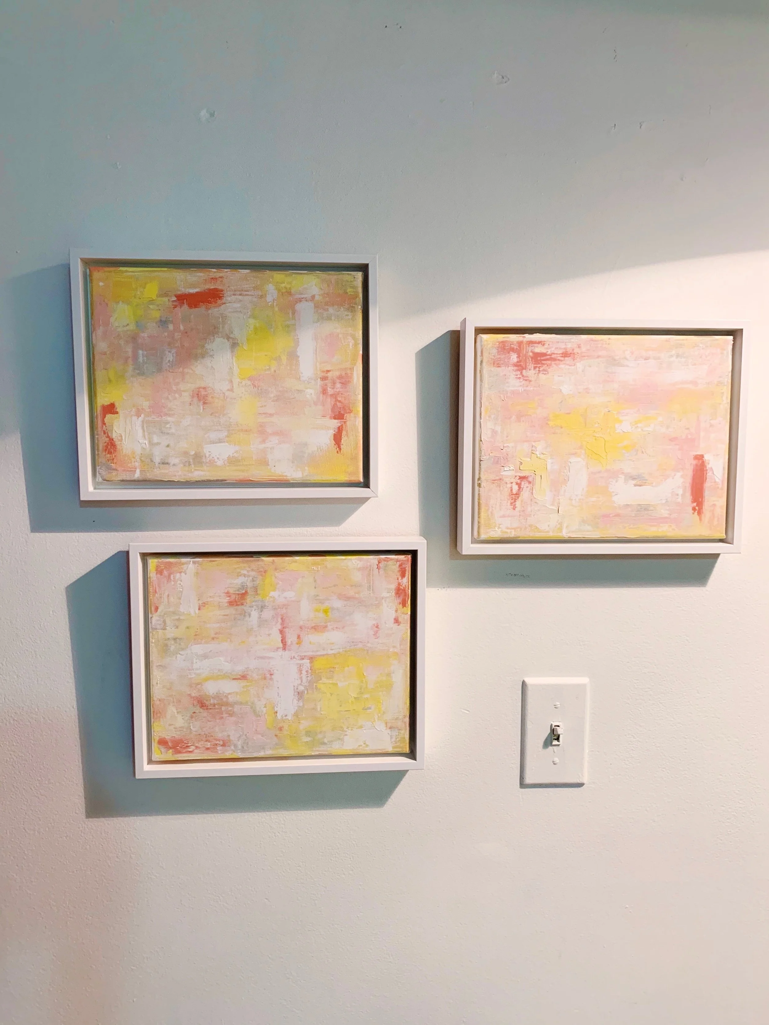 Three framed abstract paintings with pastel colors hanging on a white wall, with a power outlet and switch below.