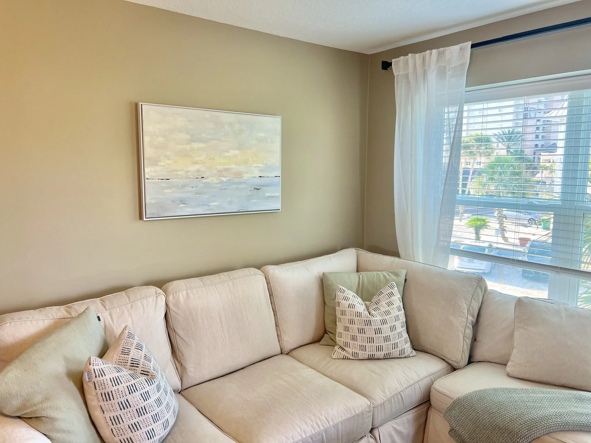 Living room area with a sectional white sofa decorated with patterned pillows, a landscape painting on the beige wall, and a large window with white curtains showing palm trees and tall buildings outside.