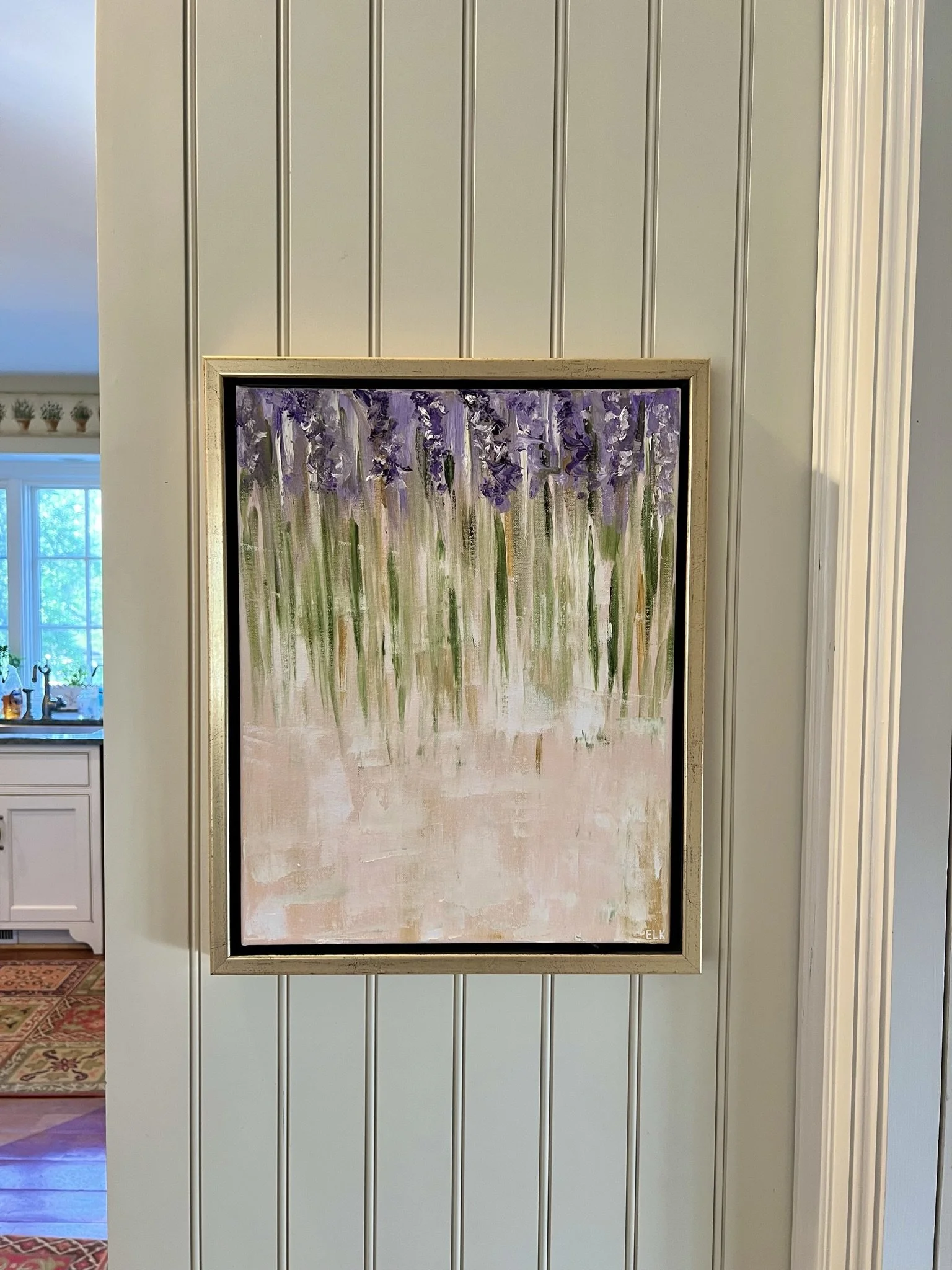 A vertical painting of purple and green flowers on a beige background, framed and hanging on a white paneled wall inside a house.