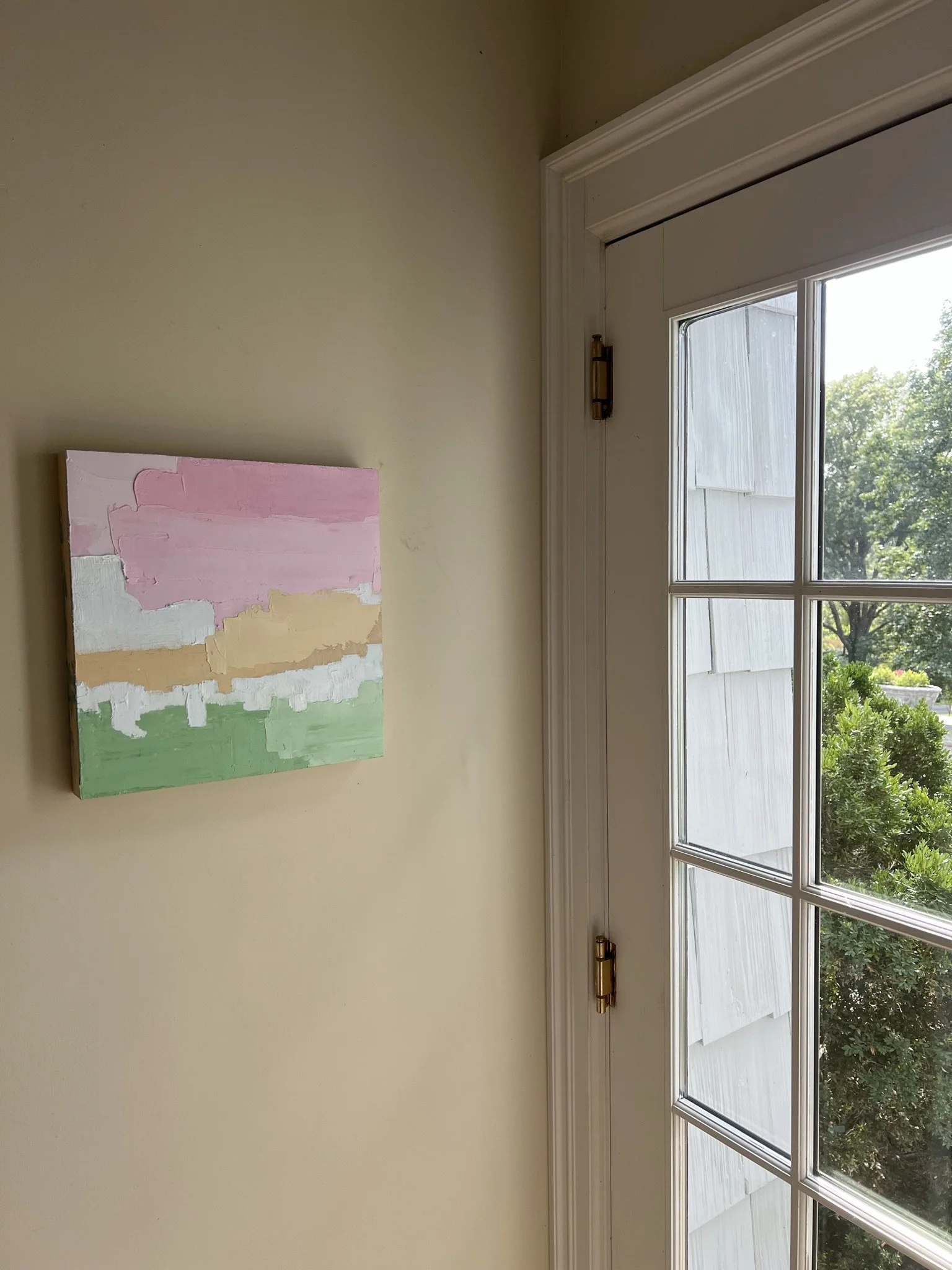 A window with white frames and divided glass panes overlooking green trees and bushes outside, next to a plain light-colored wall with a colorful abstract painting hanging.