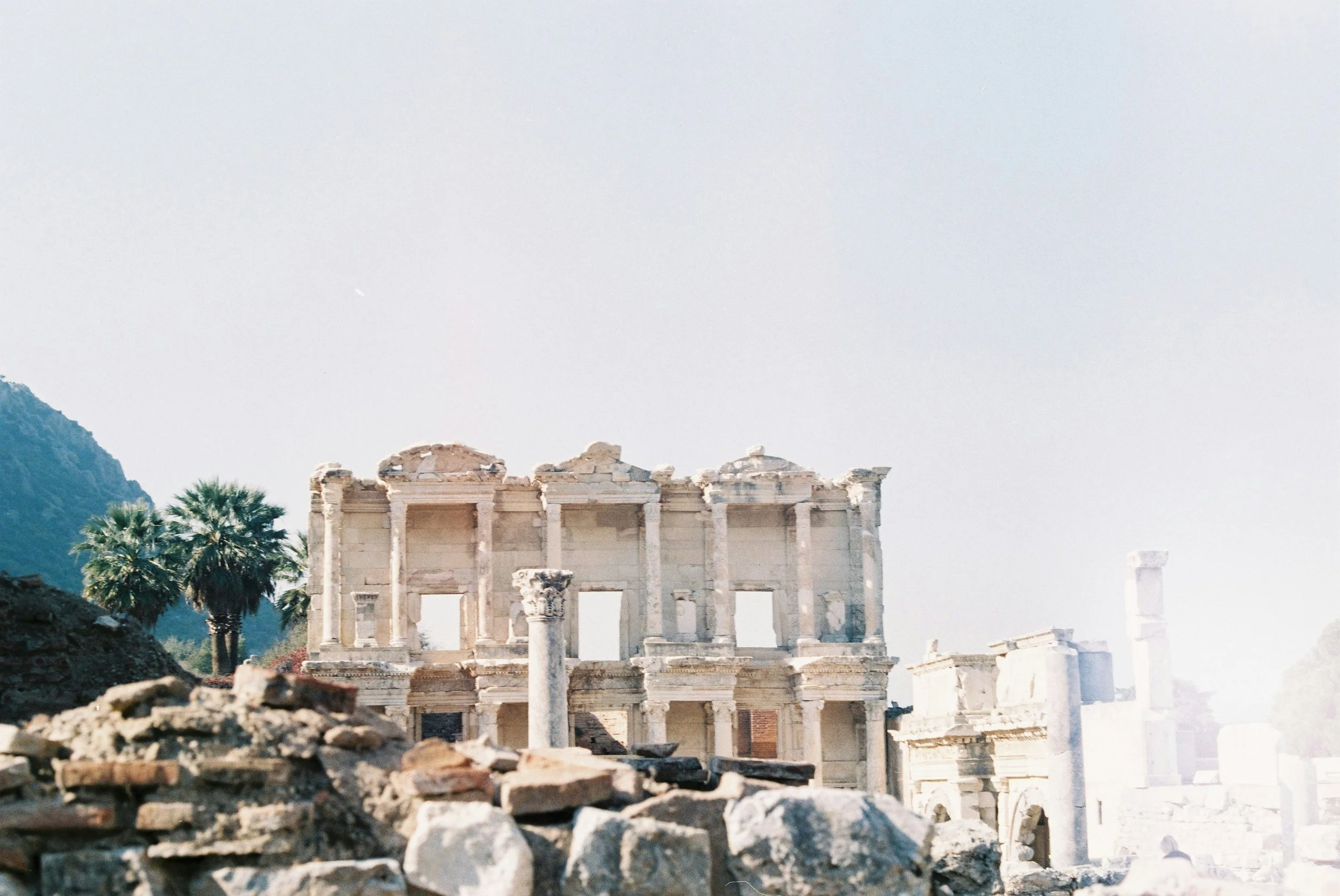 Ancient ruins of a classical building with columns, including palm trees in the background and mountainous landscape, under a clear sky.