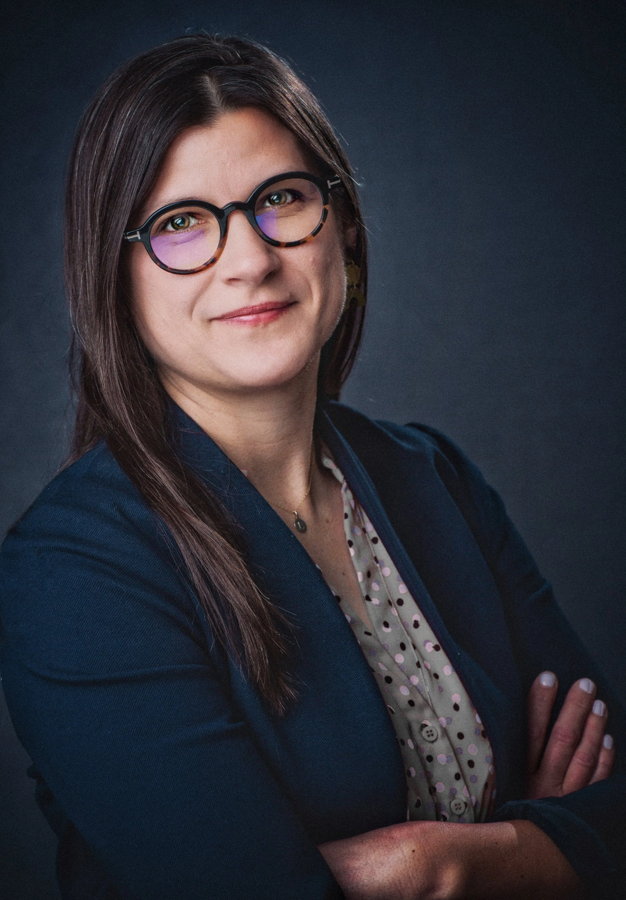 Portrait of a woman wearing black glasses with purple reflective lenses, a navy blazer, and a polka dot blouse, standing against a dark background.
