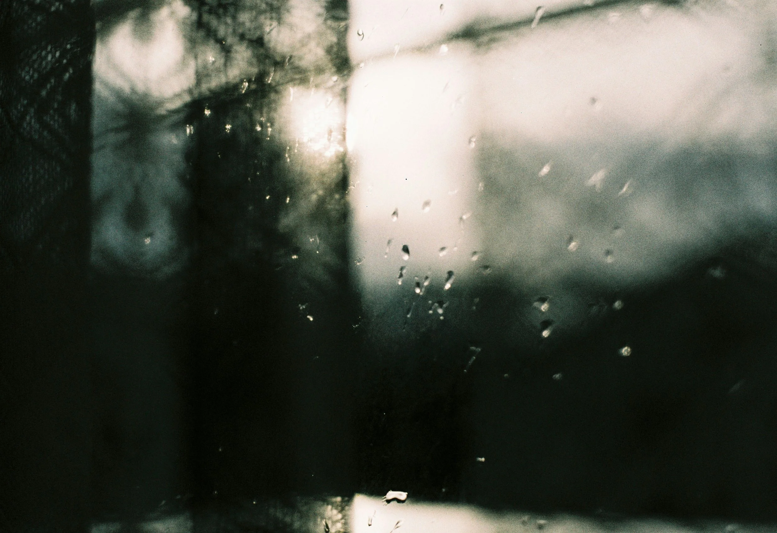 A view through a rainy window with raindrops on the glass, showing a blurry outdoors scene with overcast weather and possibly a building or structure in the distance.