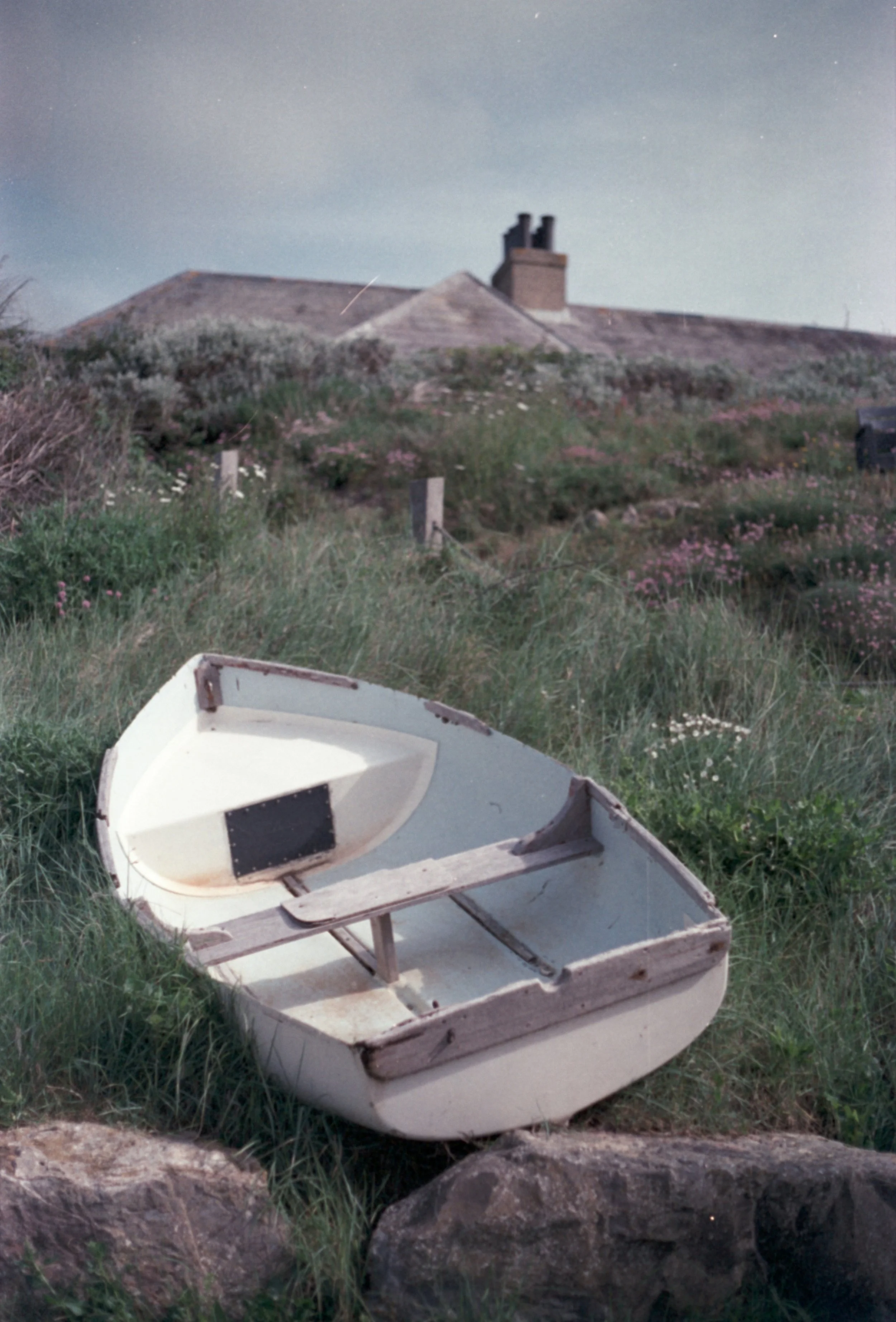 An empty, weathered wooden rowboat resting on grass and rocks near a shoreline, with a house and chimney in the background under a starry sky.