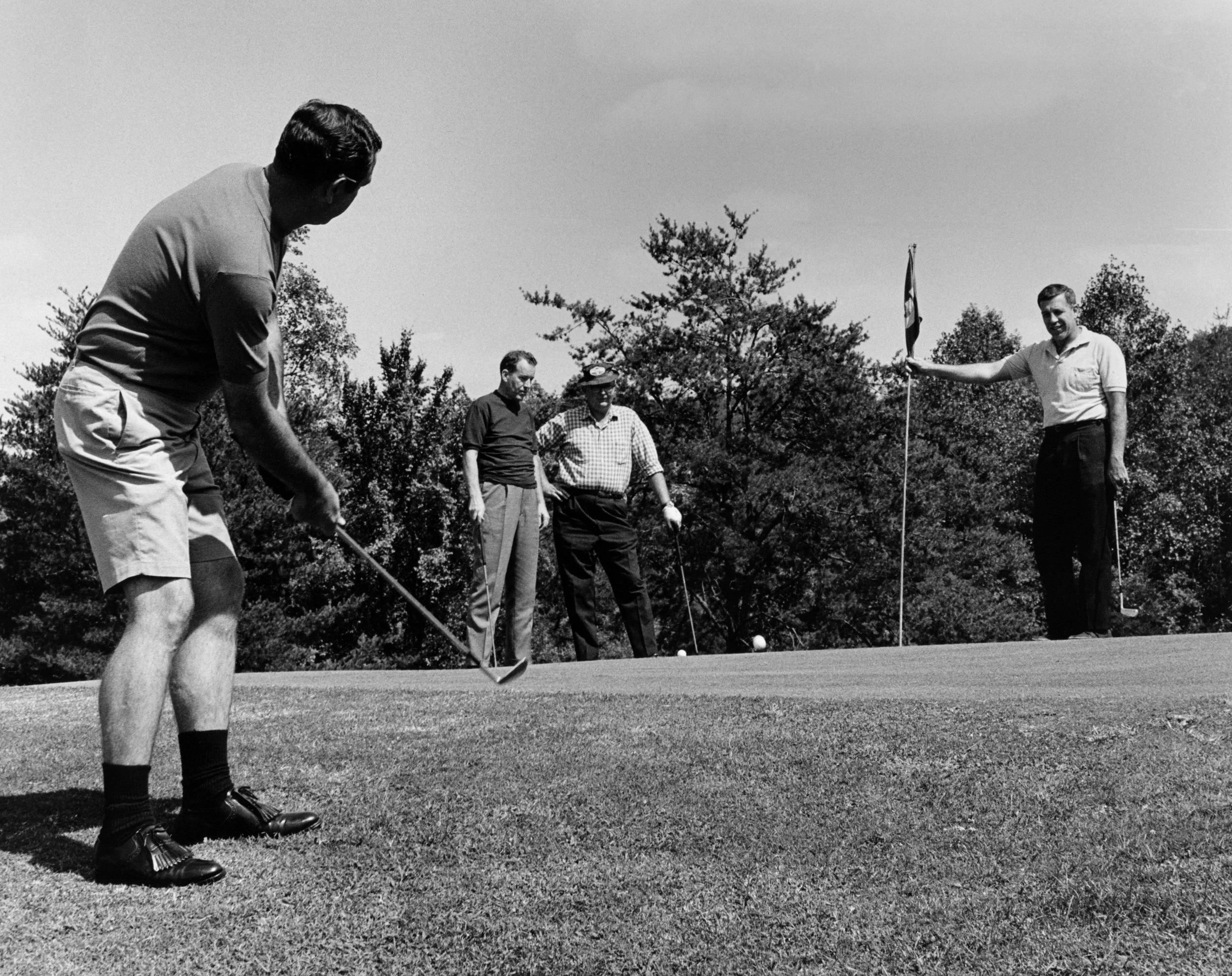 A black and white photo of four men standing on a golf course, with one man preparing to take a shot and another man holding a flag. Trees surround the area.
