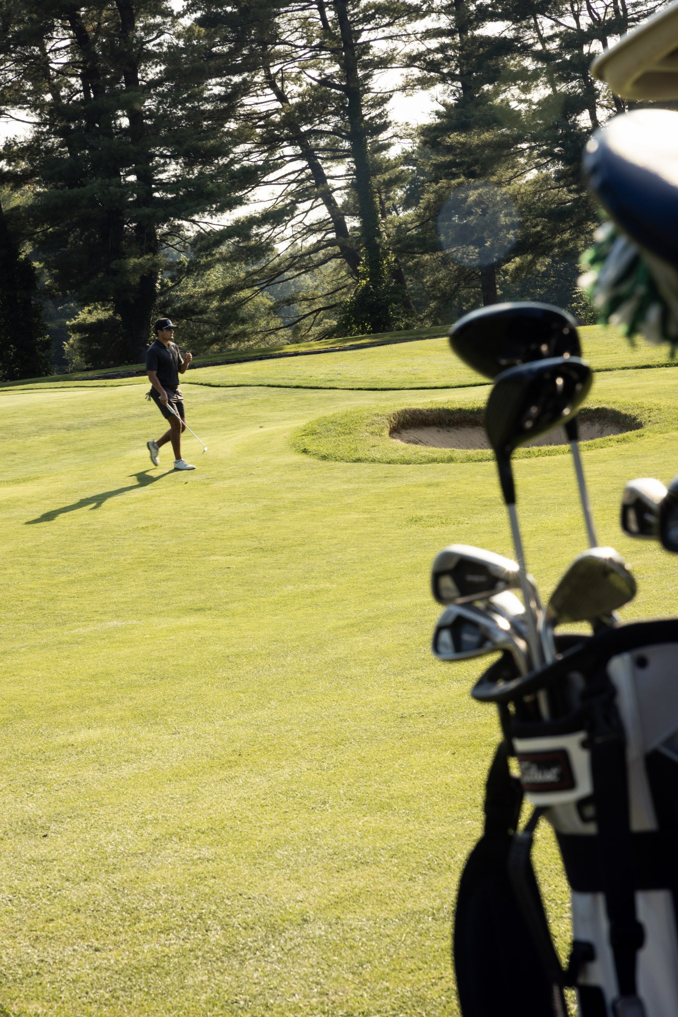 A man walking on a golf course with a golf club in his hand, surrounded by trees and sunlight, with golf clubs in the foreground.