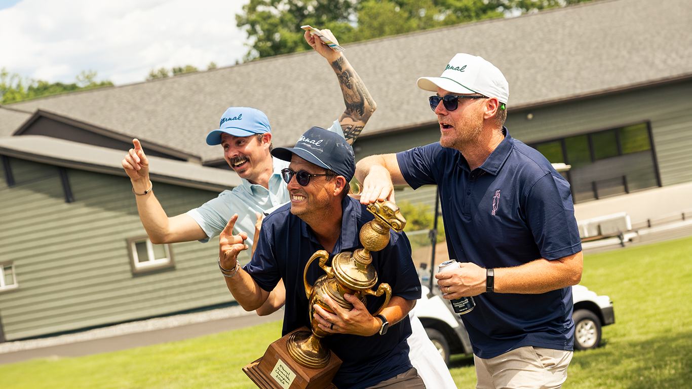 Three men celebrating on a golf course, one holding a trophy, all smiling and wearing sunglasses and golf hats.