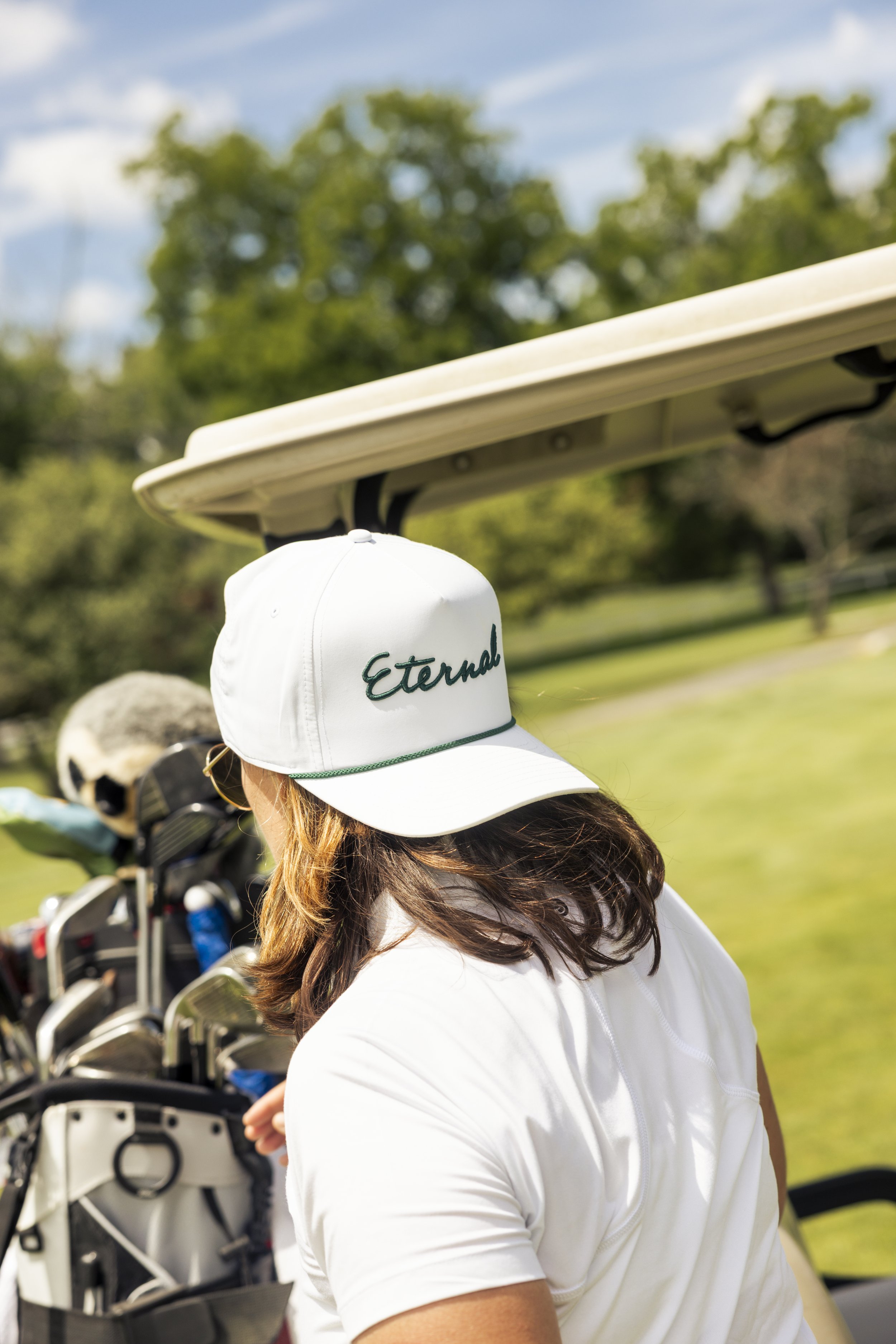 A woman with brown hair wearing a white cap with the word 'Eternal' embroidered on it, standing outdoors near a golf course with golf bags and clubs in the background.