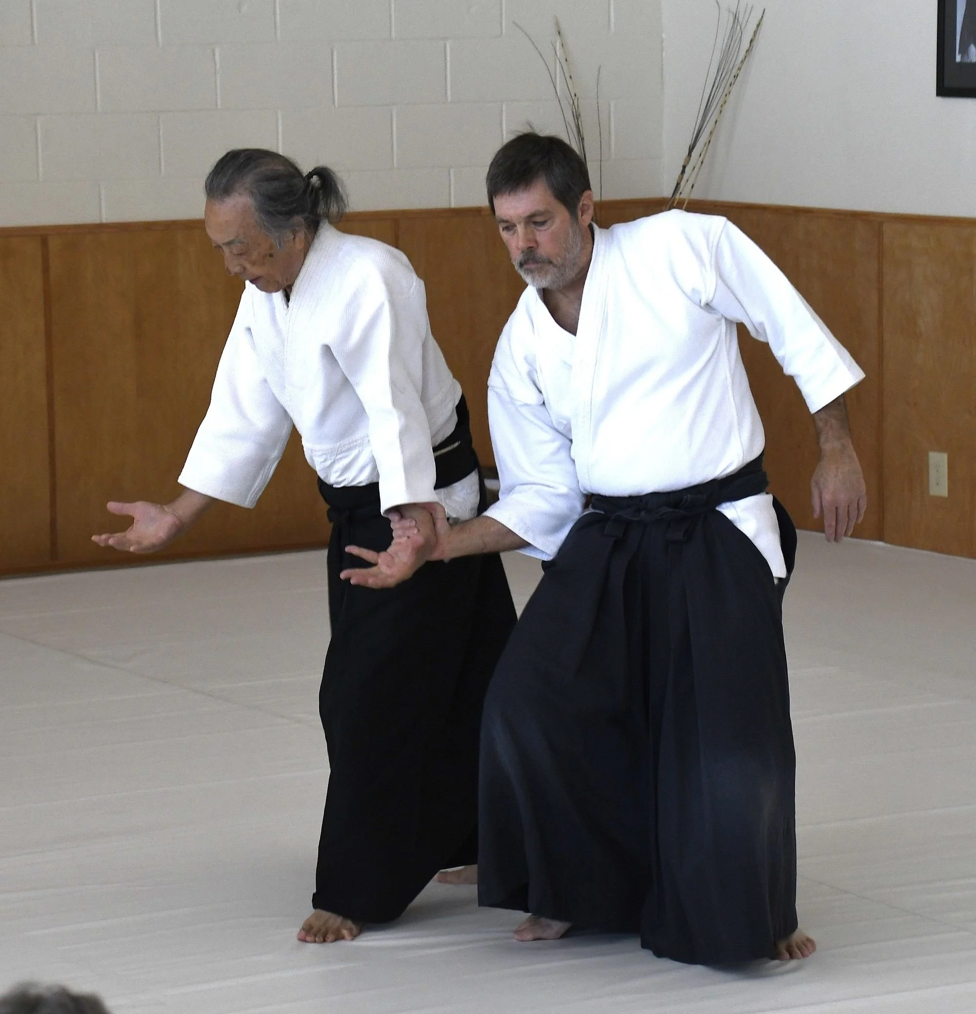 Two martial artists, a woman with gray hair and an older man with dark hair and a gray beard, practicing a joint lock technique on a light-colored mat in a dojo. Both are wearing traditional white martial arts uniforms.