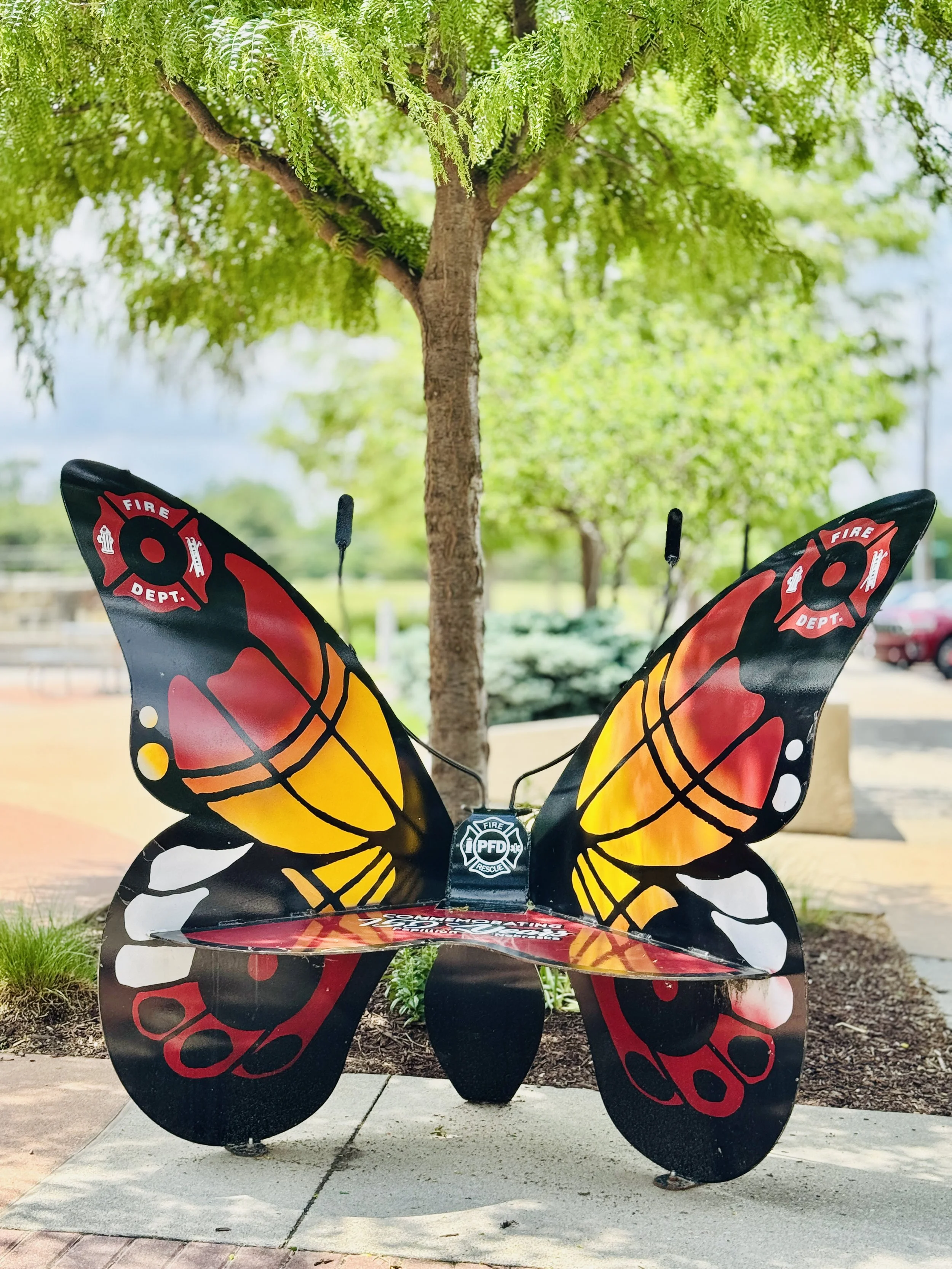 Colorful butterfly-shaped public safety memorial sculpture with firefighter symbols, placed outdoors on a concrete walkway with grass and trees in the background.