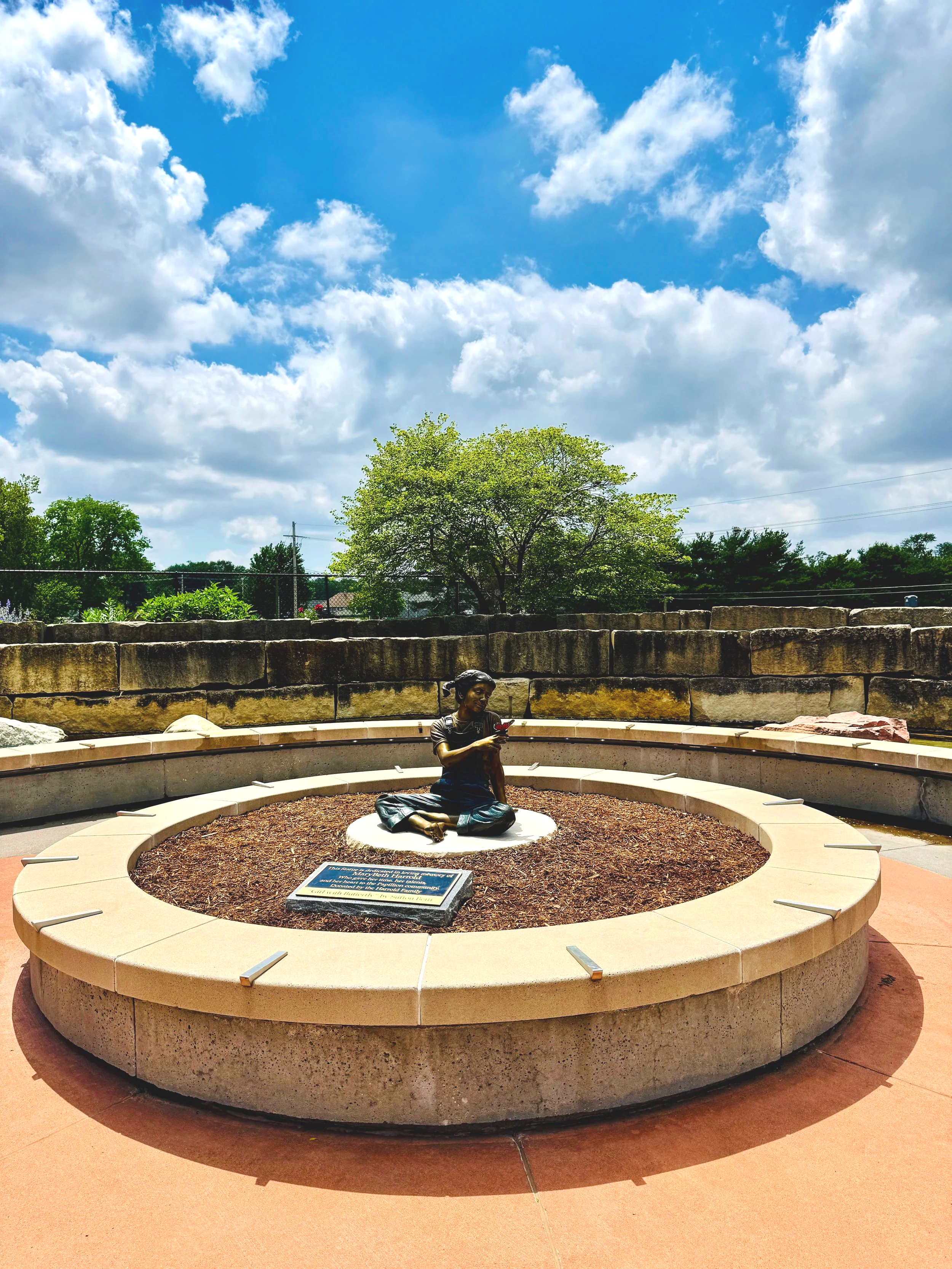 Bronze statue of a woman sitting cross-legged and reading a book, situated on a circular stone platform with a plaque, surrounded by a garden with trees and a bright blue sky with scattered clouds in the background.