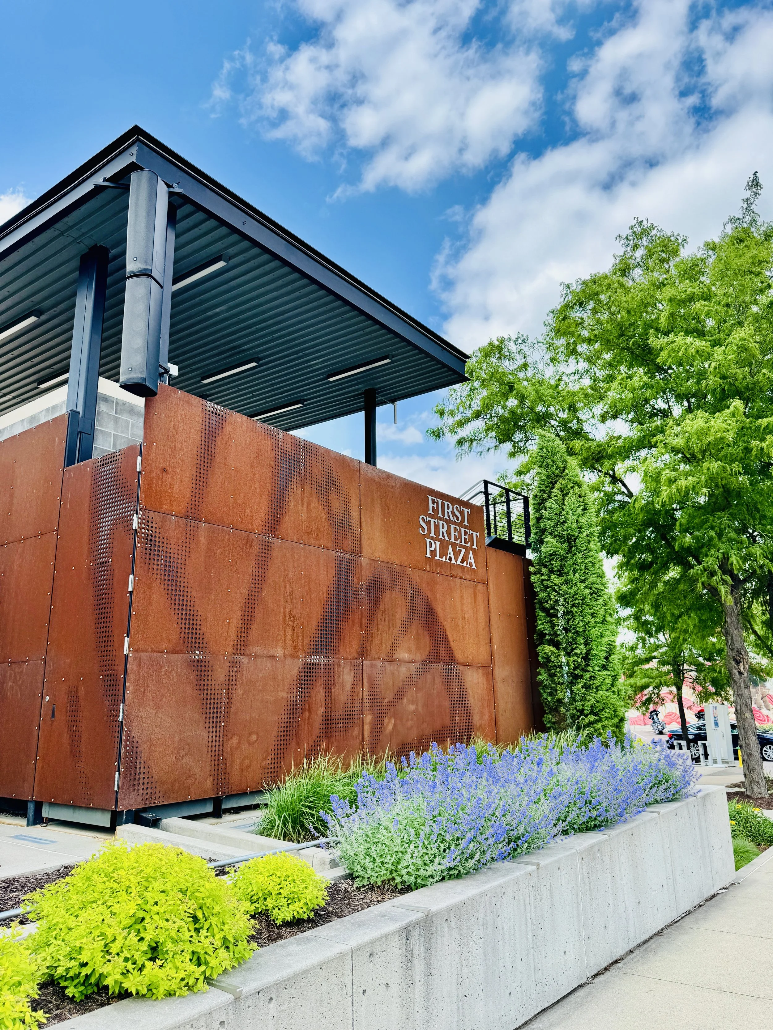 Exterior view of a modern building with a rust-colored metal facade, green trees, and a garden bed with purple and yellow flowers, under a blue sky with scattered clouds. Sign on the building reads "First Street Plaza."