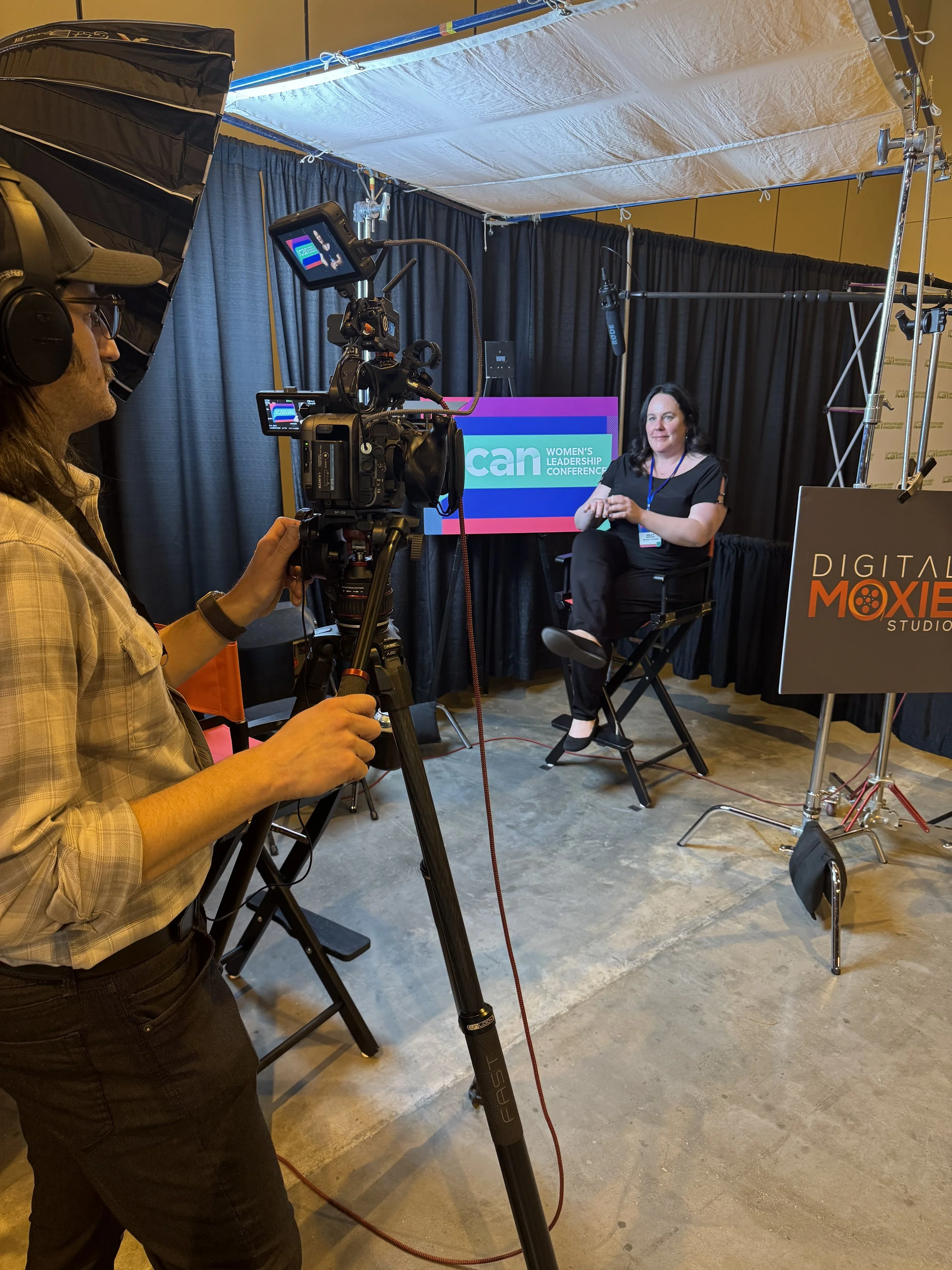 A woman being filmed in a video interview setup with a camera operator in front, professional lighting, a backdrop with curtains, and a screen displaying 'CAN Women's Leadership Conference' behind her.