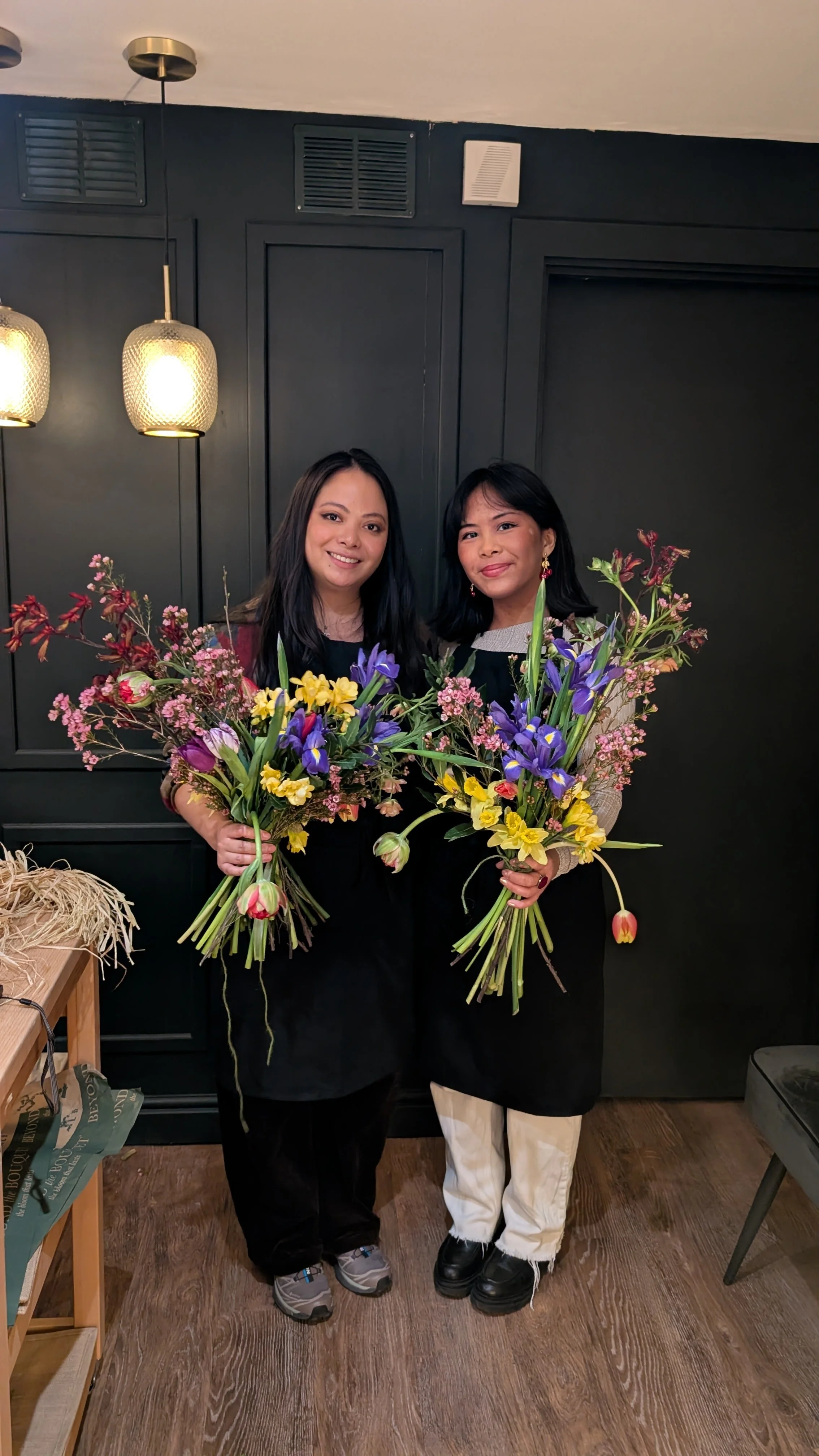 Two women standing indoors holding large bouquets of colorful flowers, smiling at the camera, against a dark wall background with warm lighting.