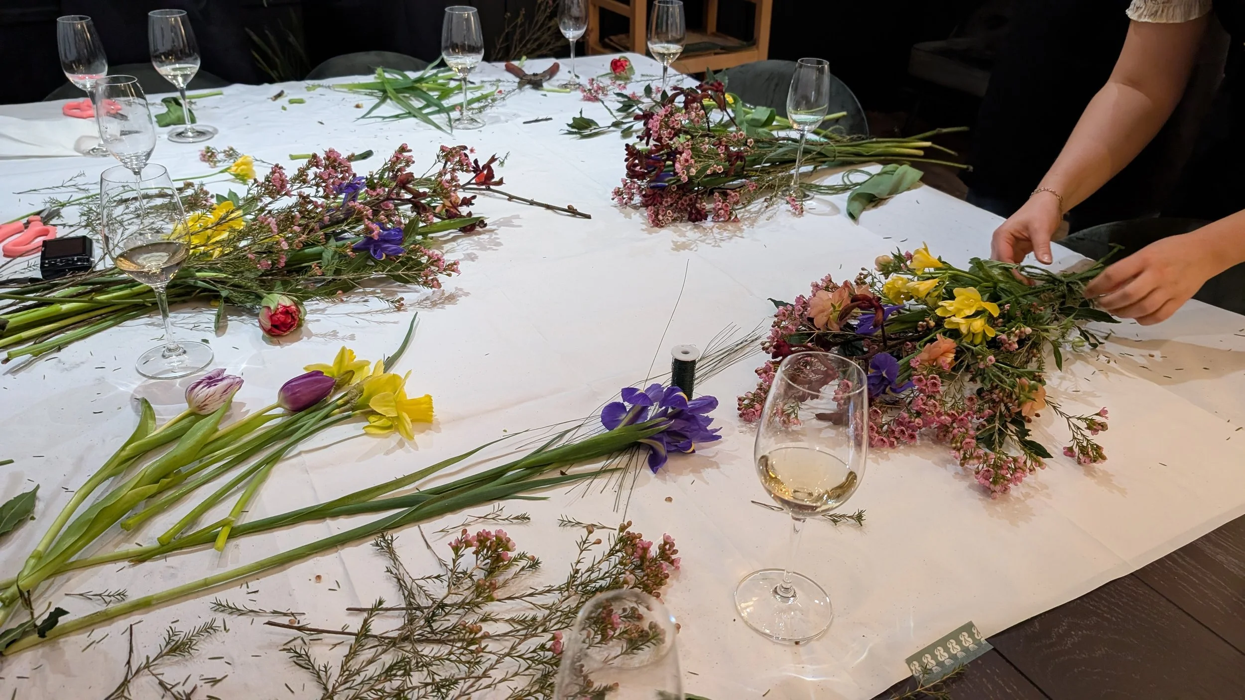 A bouquet of various flowers being arranged on a table with several wine glasses and scattered flowers, indicating flower arranging or floral decoration work.