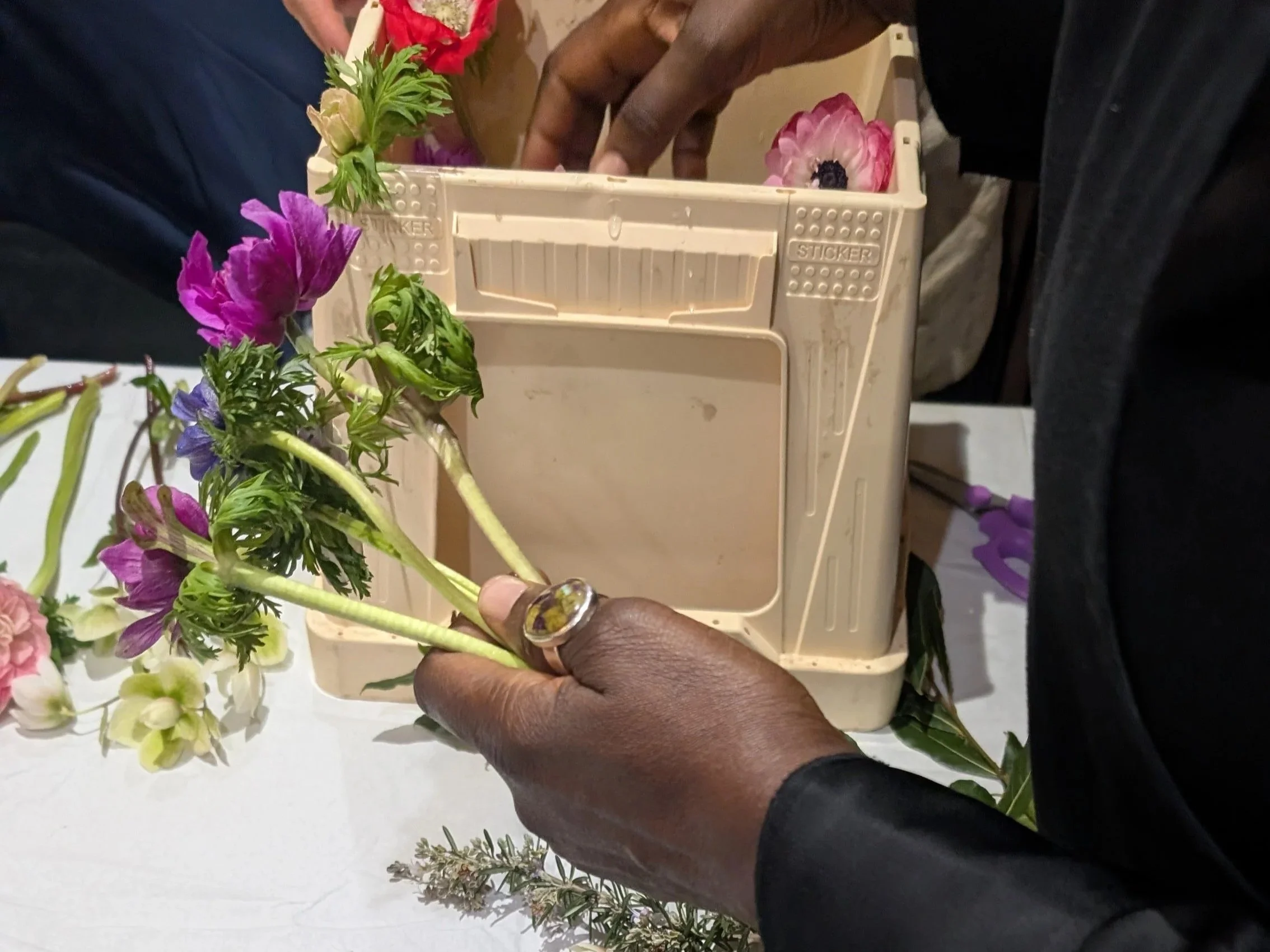Close-up of hands arranging colorful artificial flowers in a small plastic basket, with some additional floral pieces on a white surface nearby.