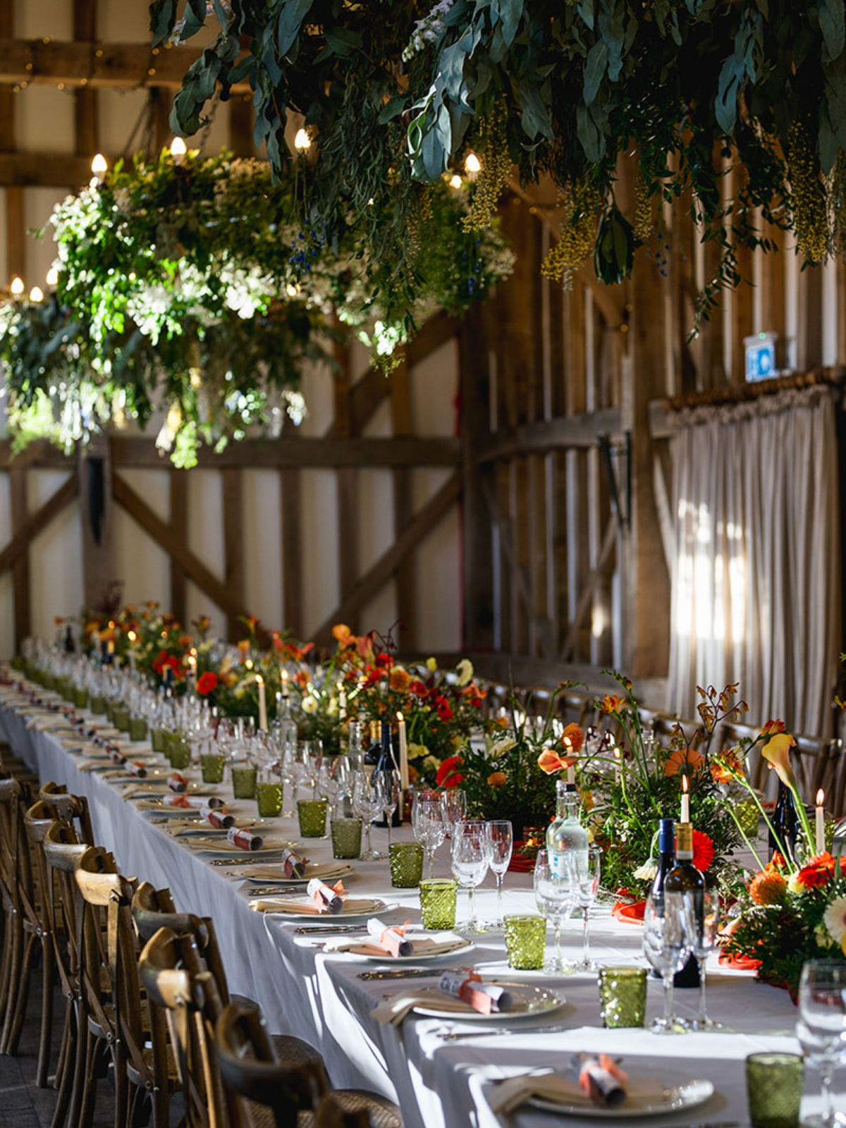 A long banquet table decorated with flowers, candles, and glasses, set in a rustic barn with wooden walls and hanging green foliage.