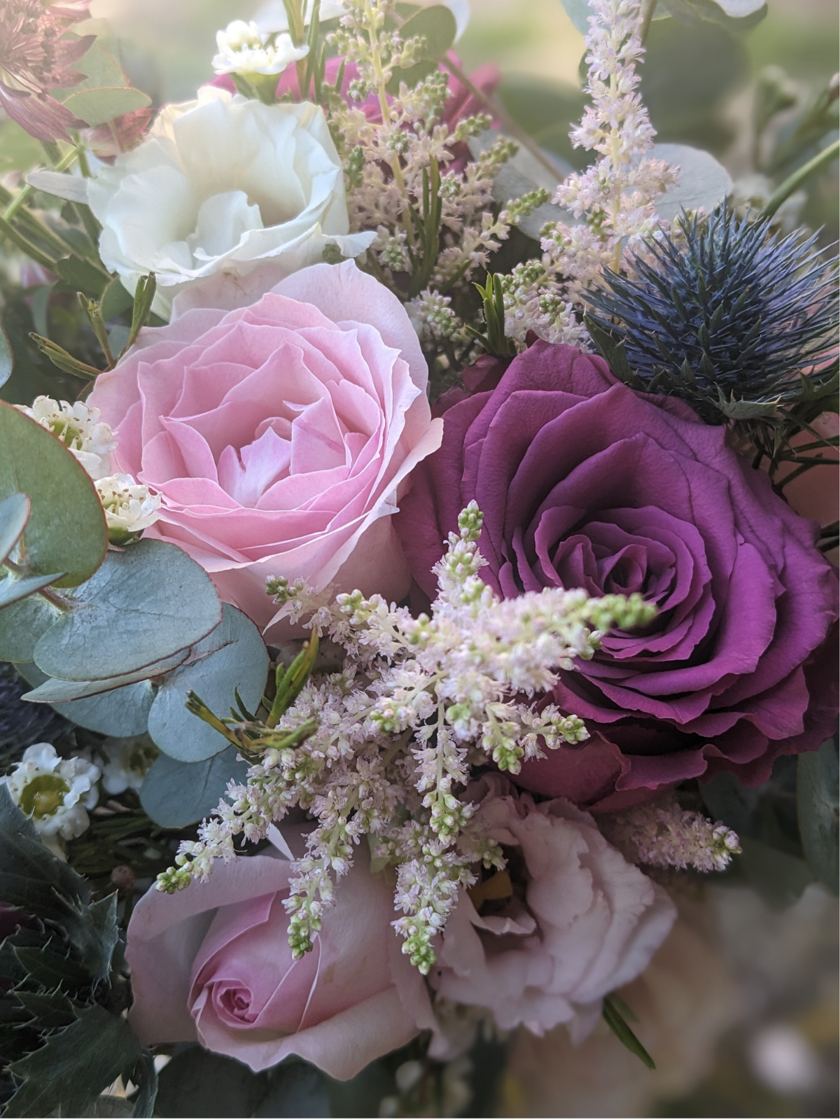 A close-up of a floral arrangement with pink and purple roses, white lisianthus, and smaller white and pink flowers, along with green eucalyptus leaves and a blue thistle flower.