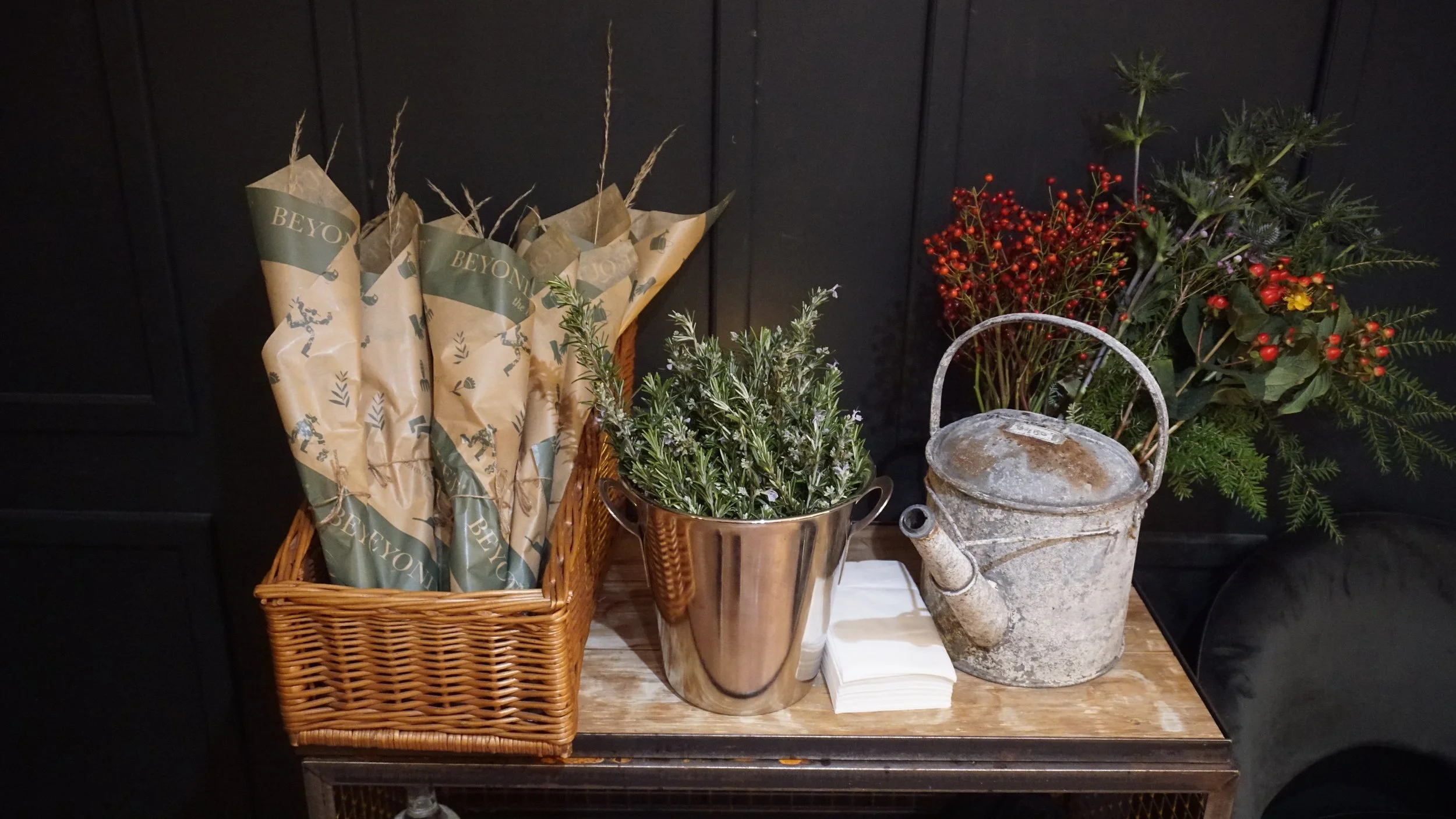 A display of potted plants, dried flowers, a wicker basket with paper-wrapped items, a metal watering can, and white napkins, arranged on a wooden surface against a dark background.