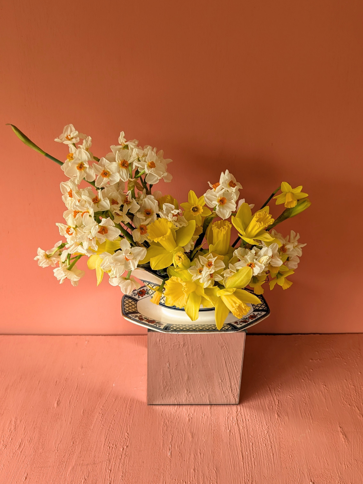 A floral arrangement with white and yellow flowers in a decorative bowl on a square mirror on a pink pedestal against a pink wall.