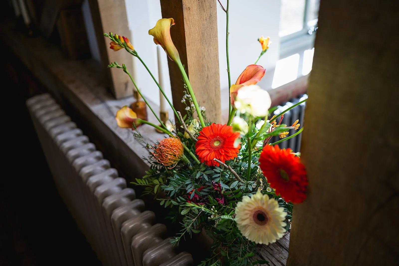 Colorful flower arrangement with orange gerbera daisies, a peach-colored calla lily, white and yellow ranunculus, and various greenery, placed on a windowsill next to a radiator.