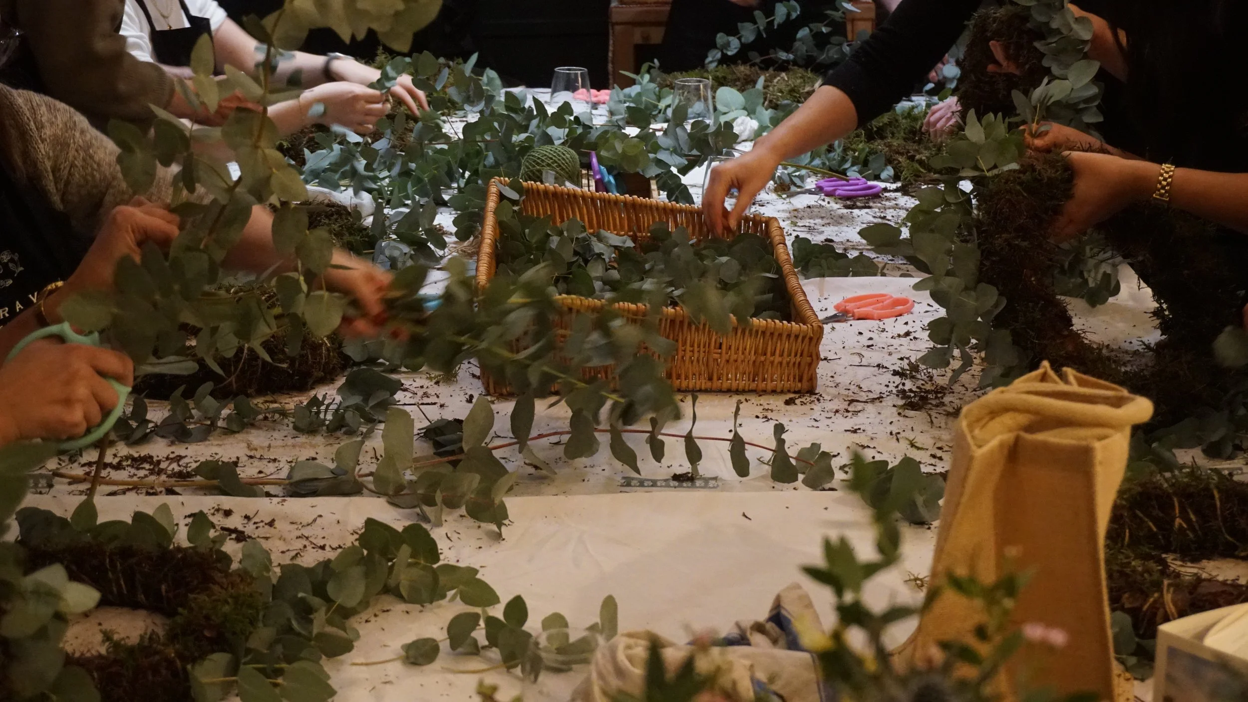 People gathered around a table creating floral arrangements with eucalyptus and other greenery, using scissors, with baskets and supplies on the table.
