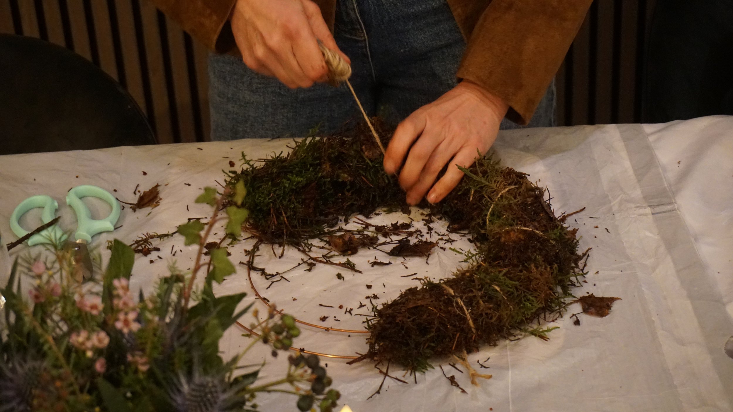 Person dressed in a brown jacket and gray pants arranging a nativity scene with moss, plants, and figurines on a white surface.