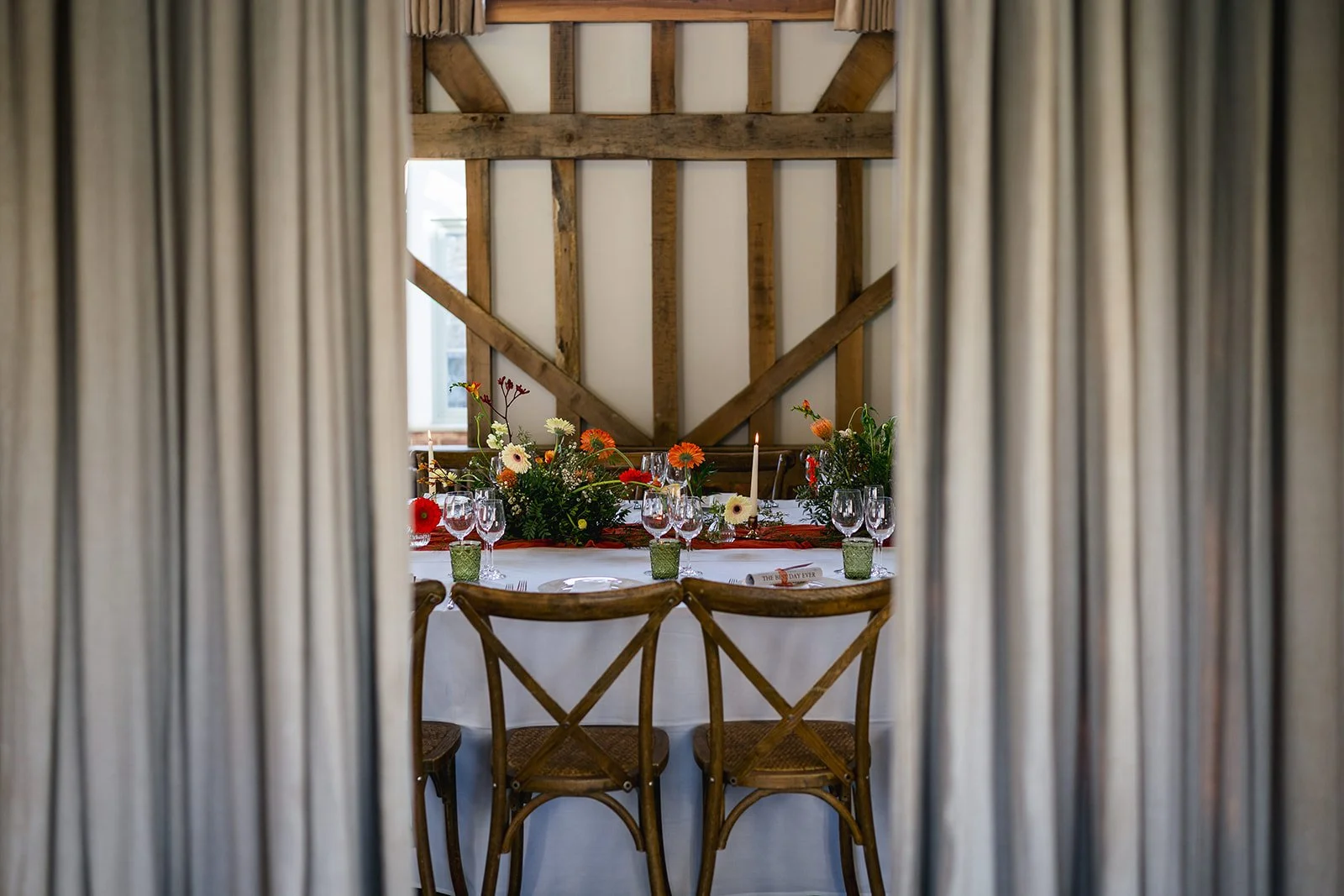 A dining table set for a celebration or event, with a floral centerpiece, multiple wine glasses, candles, and place cards, viewed through a doorway with curtains on either side in a rustic room with wooden beams.