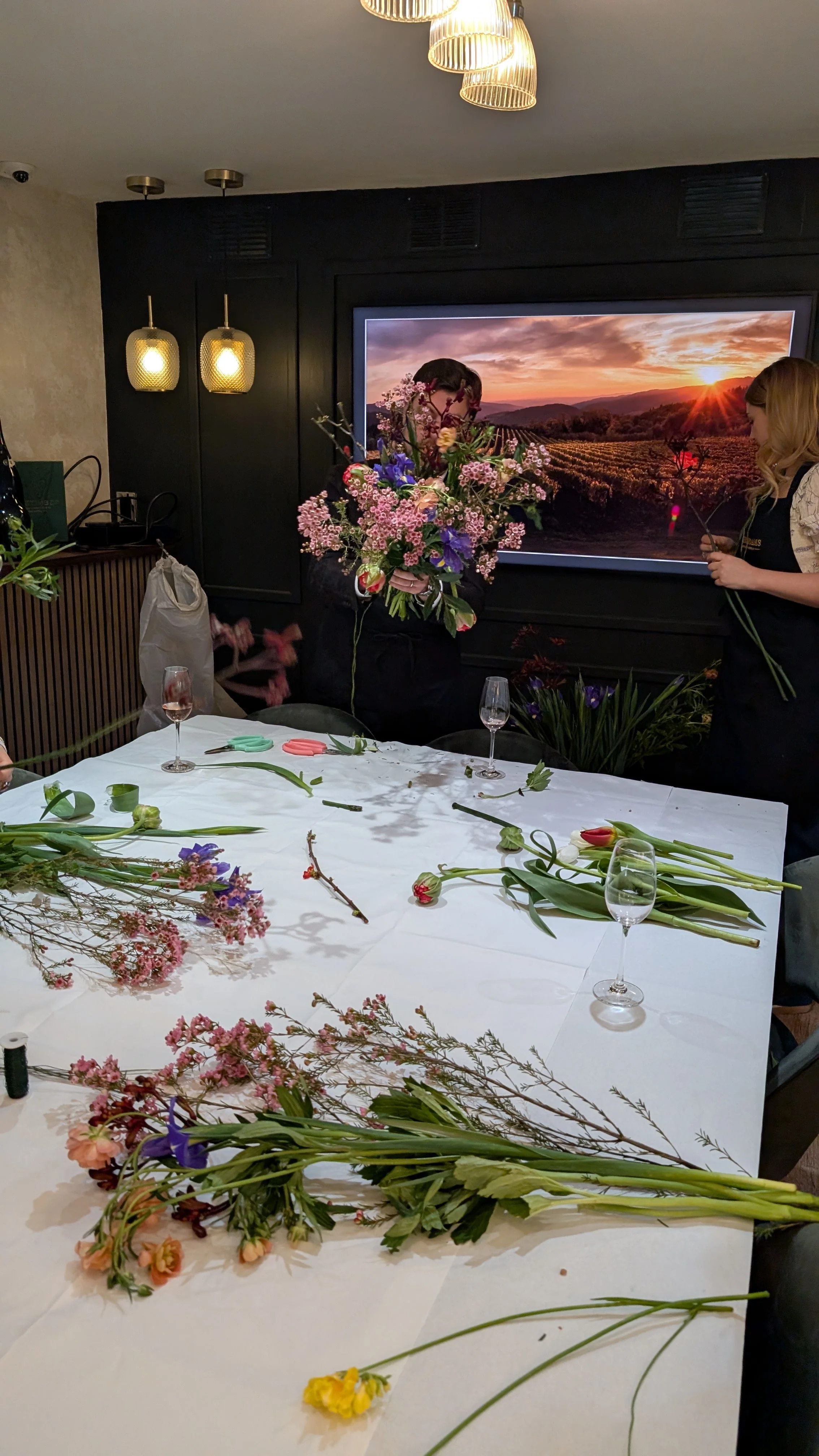 People preparing flower arrangements in a room with elegant lighting and a large screen displaying a scenic sunset over a vineyard.