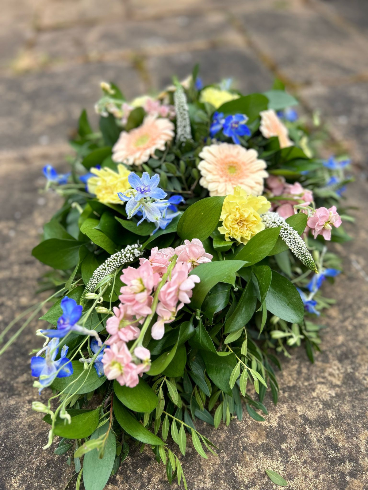 A floral arrangement with pastel pink, yellow, and blue flowers, green leaves, and white accents laid on a stone surface.