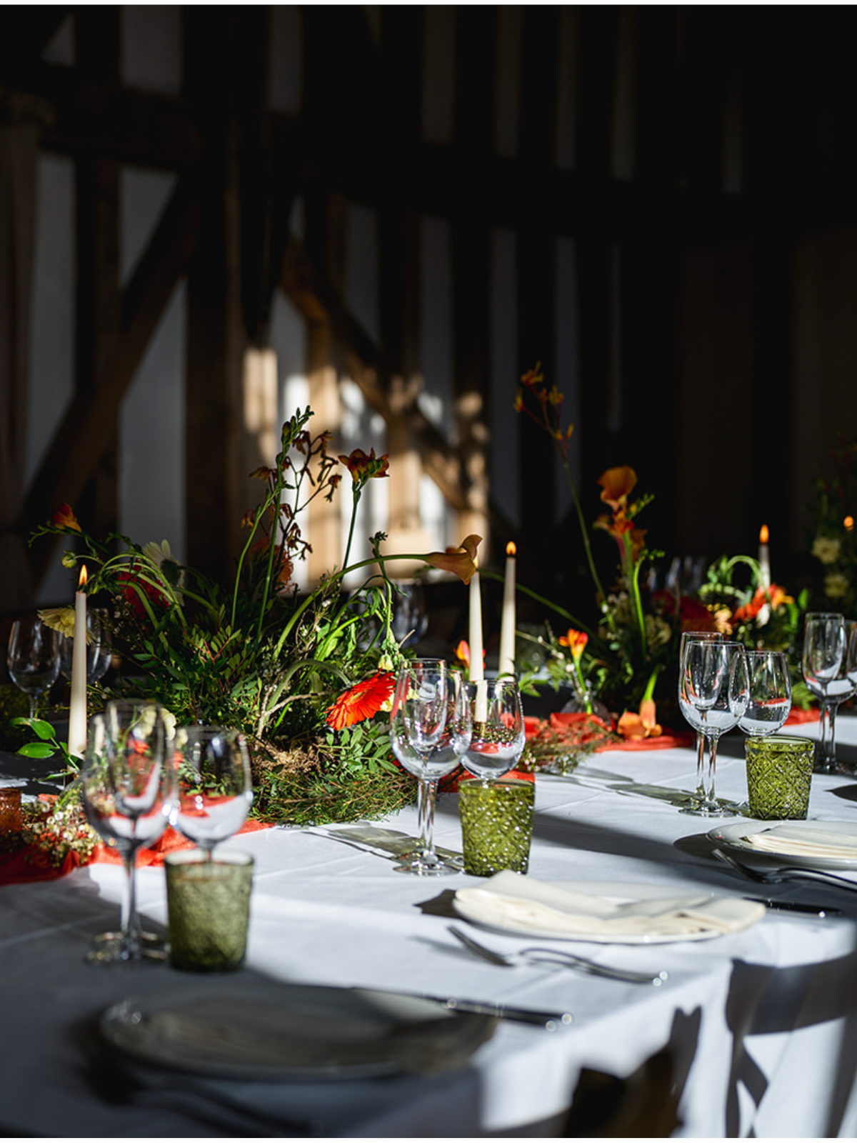 Elegant banquet table set for a formal event, with floral centerpieces, candles, wine glasses, and tableware in a sunlit room with wooden beams.