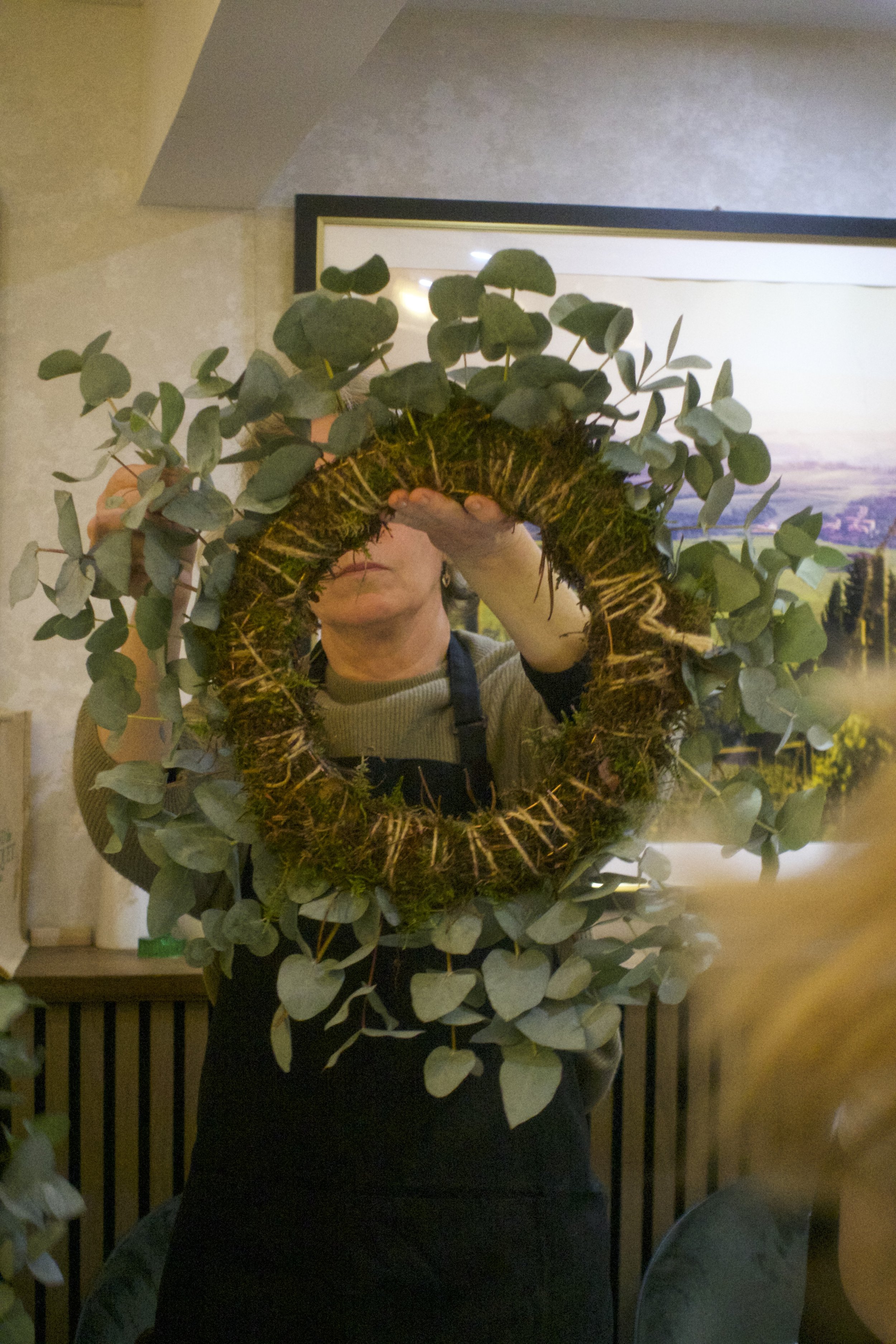 A person is holding a round moss and eucalyptus leaf wreath in a room with a framed picture of a sunset or landscape on the wall. The person's face is partially visible, and they are wearing a beige sweater, with their other arm in a black sleeve. Th