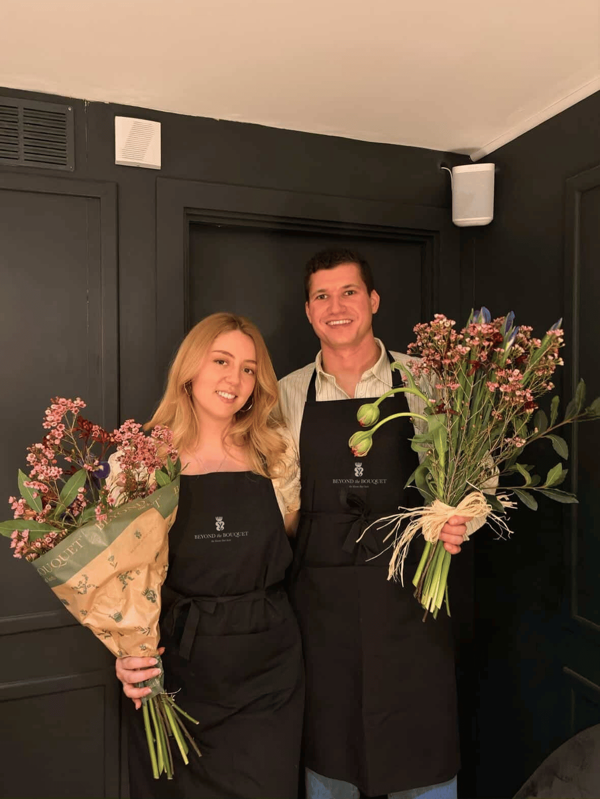 A man and woman standing together indoors, each holding a bouquet of flowers, both wearing black aprons with 'BEYOND THE BOUQUET' logo, smiling, with a dark wall background.