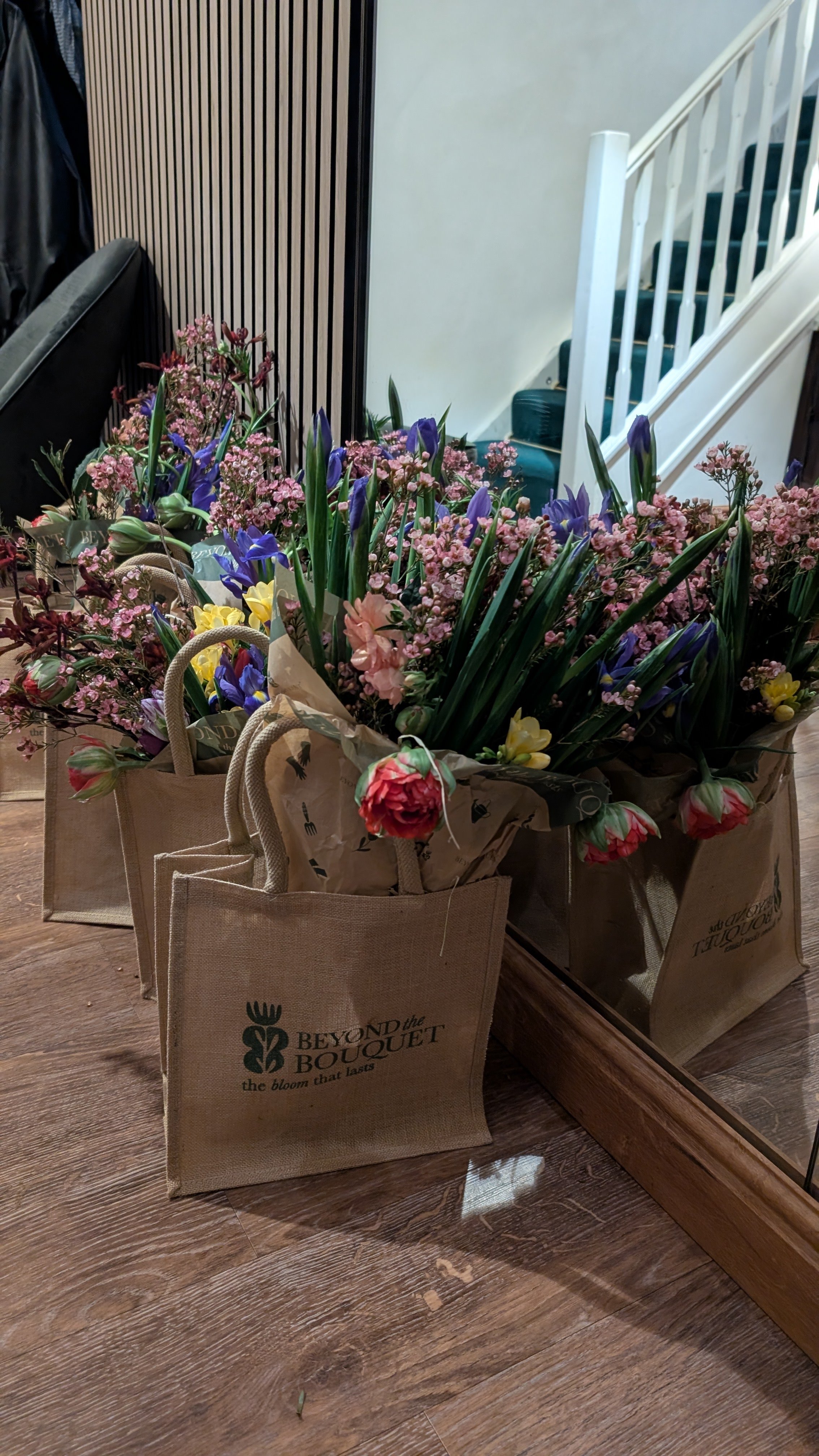 Several paper shopping bags filled with a colorful assortment of flowers, including purple irises, pink flowers, and yellow blooms, placed on a hardwood floor in front of a mirror.