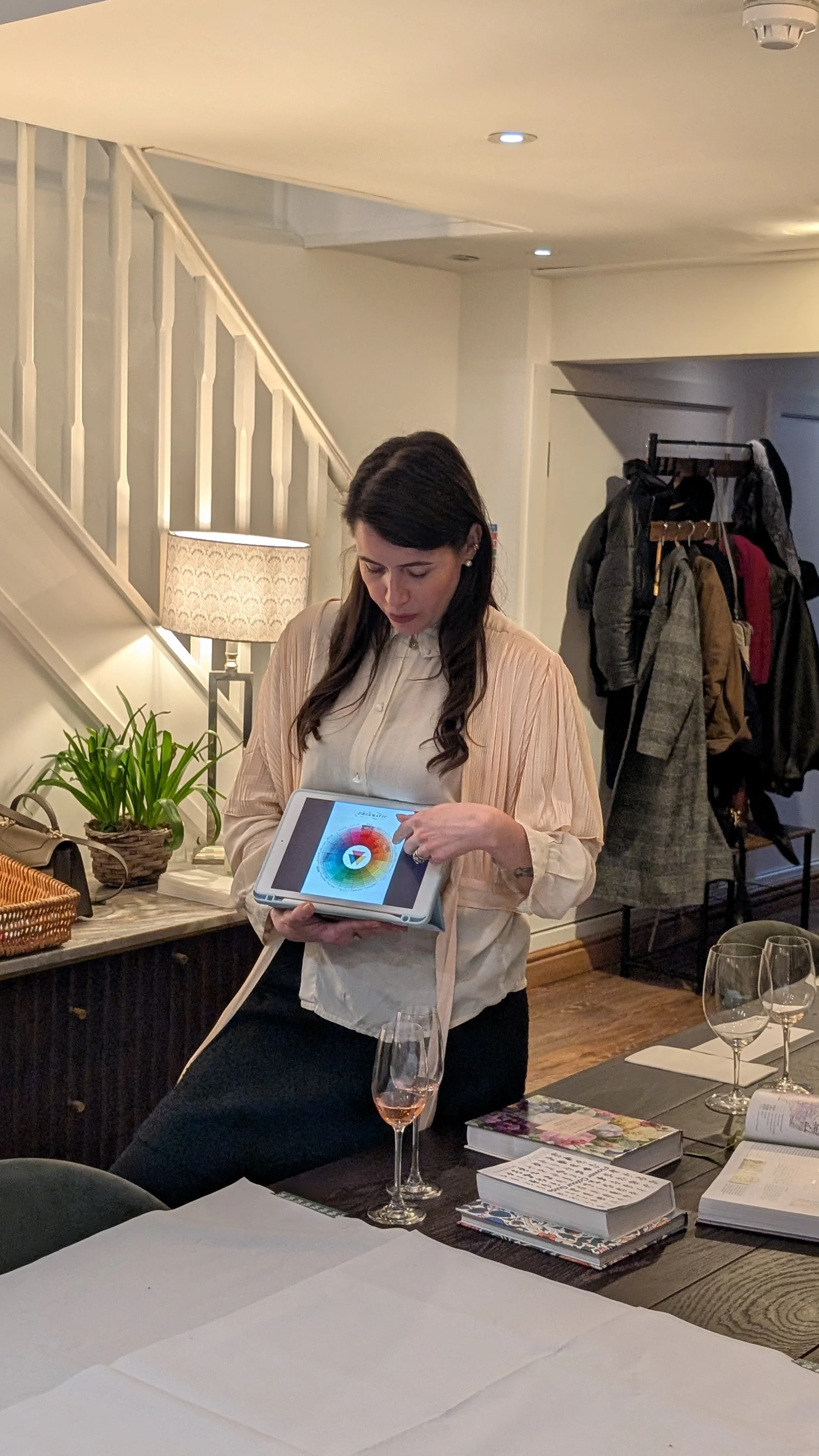 A woman standing at a table with wine glasses, using a tablet. There are books and papers on the table, a coat rack with jackets in the background, and a lamp and plants nearby.