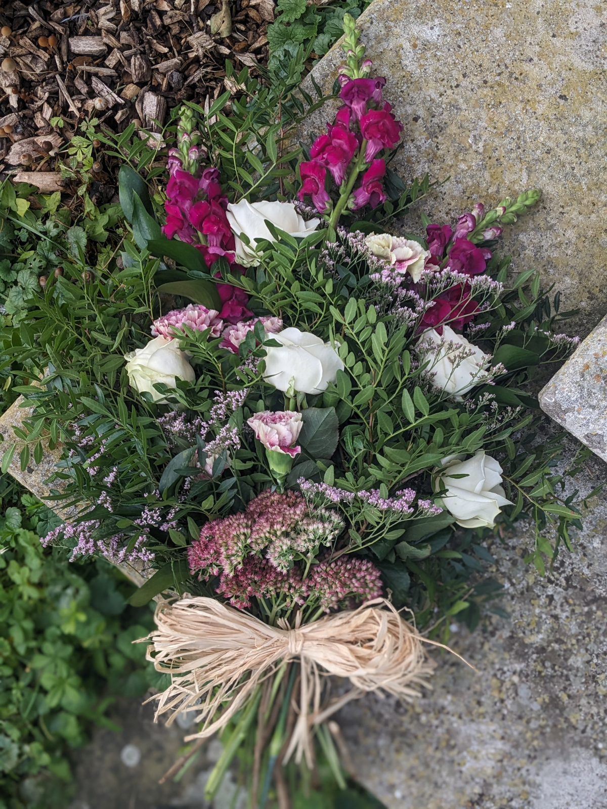A bouquet of mixed flowers including white roses, pink snapdragons, and purple heather, tied with a raffia ribbon, placed on a stone surface.