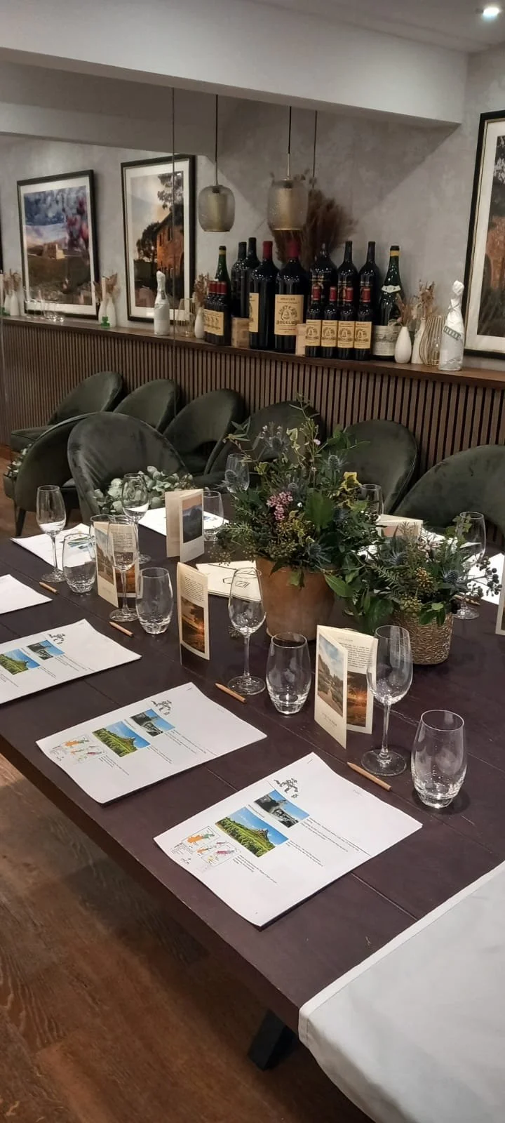 A dining table set for a meal with menus, glasses, and a floral centerpiece in a cozy restaurant with framed pictures, wine bottles, and decorative vases in the background.