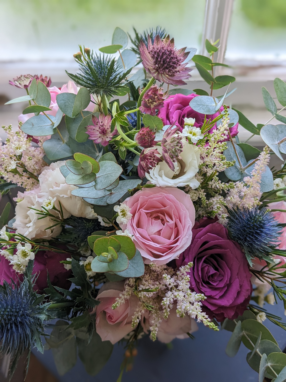 A colorful bouquet of various flowers including pink and purple roses, white lisianthus, blue eryngium, and green foliage.