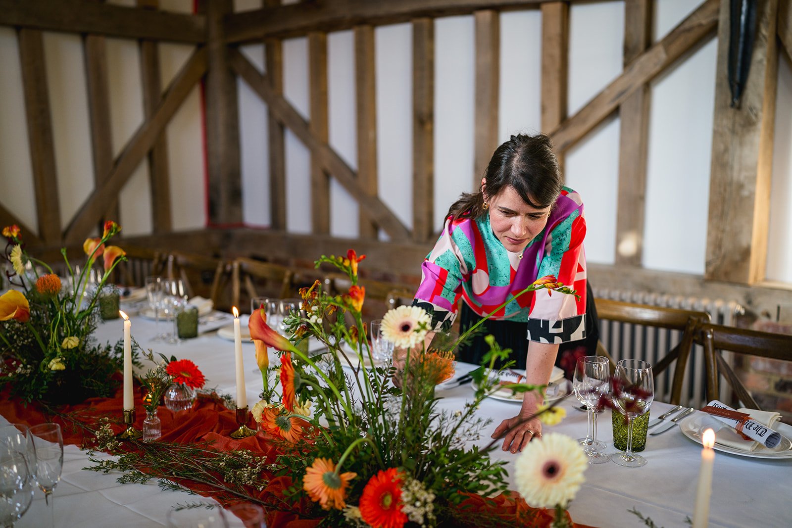 A woman setting a table decorated with colorful flowers and candles in a rustic wooden room.