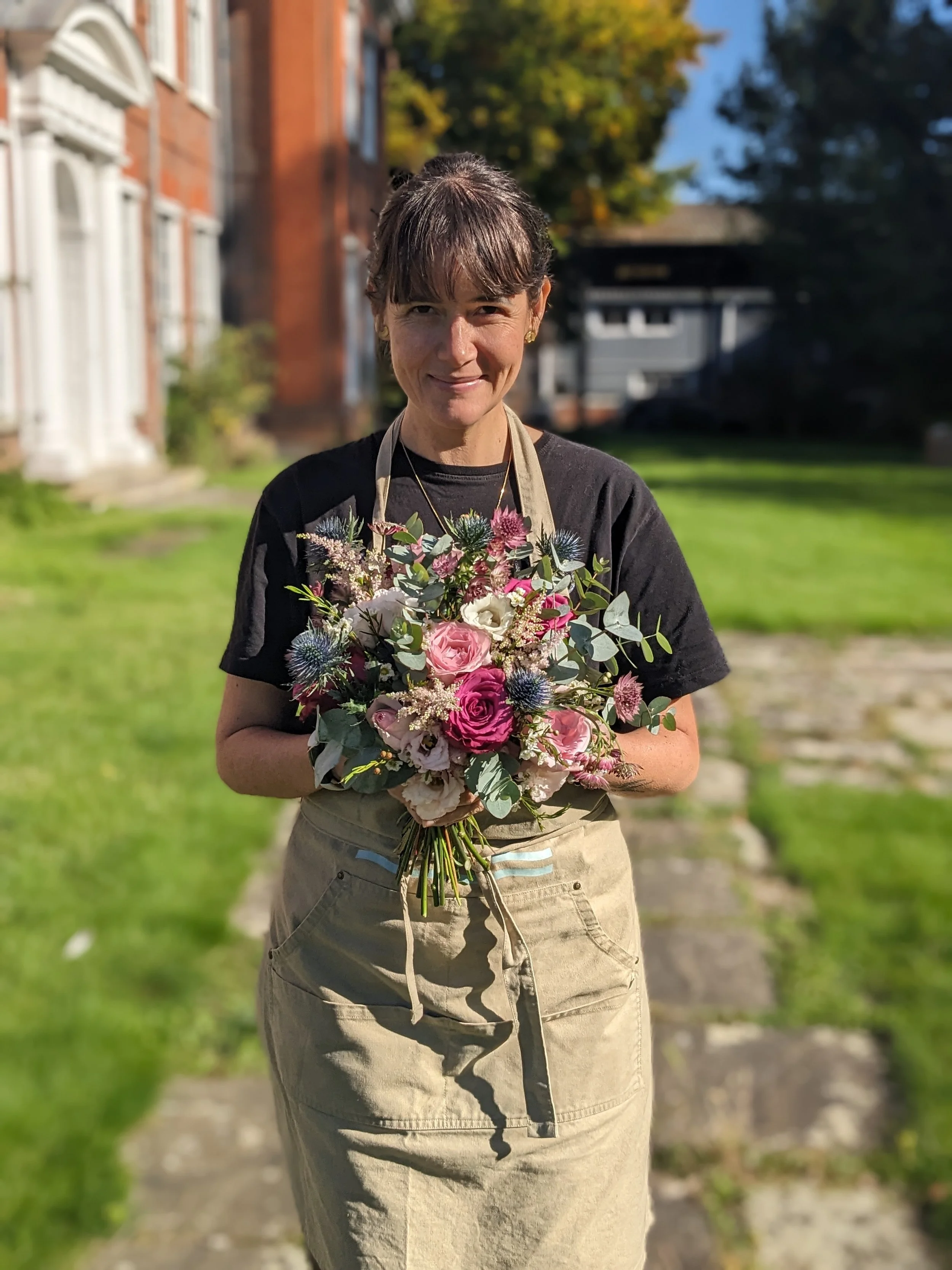 A woman standing outside on a sunny day, holding a bouquet of flowers, wearing a black T-shirt and tan cargo shorts, with trees and buildings in the background.