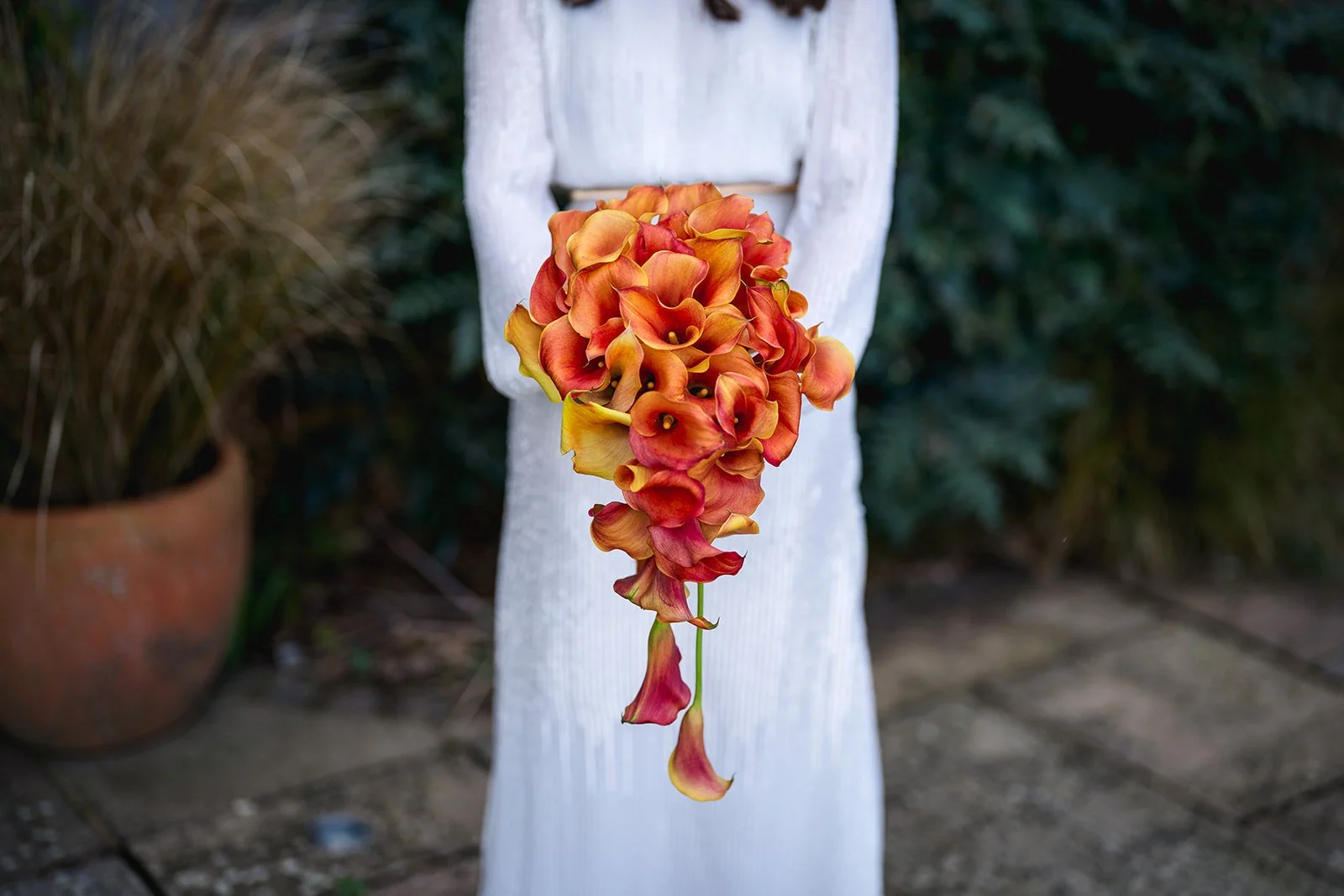 A bunch of orange and yellow flowers hanging from a white, vertical standing object with a blurred background of greenery and a pot.