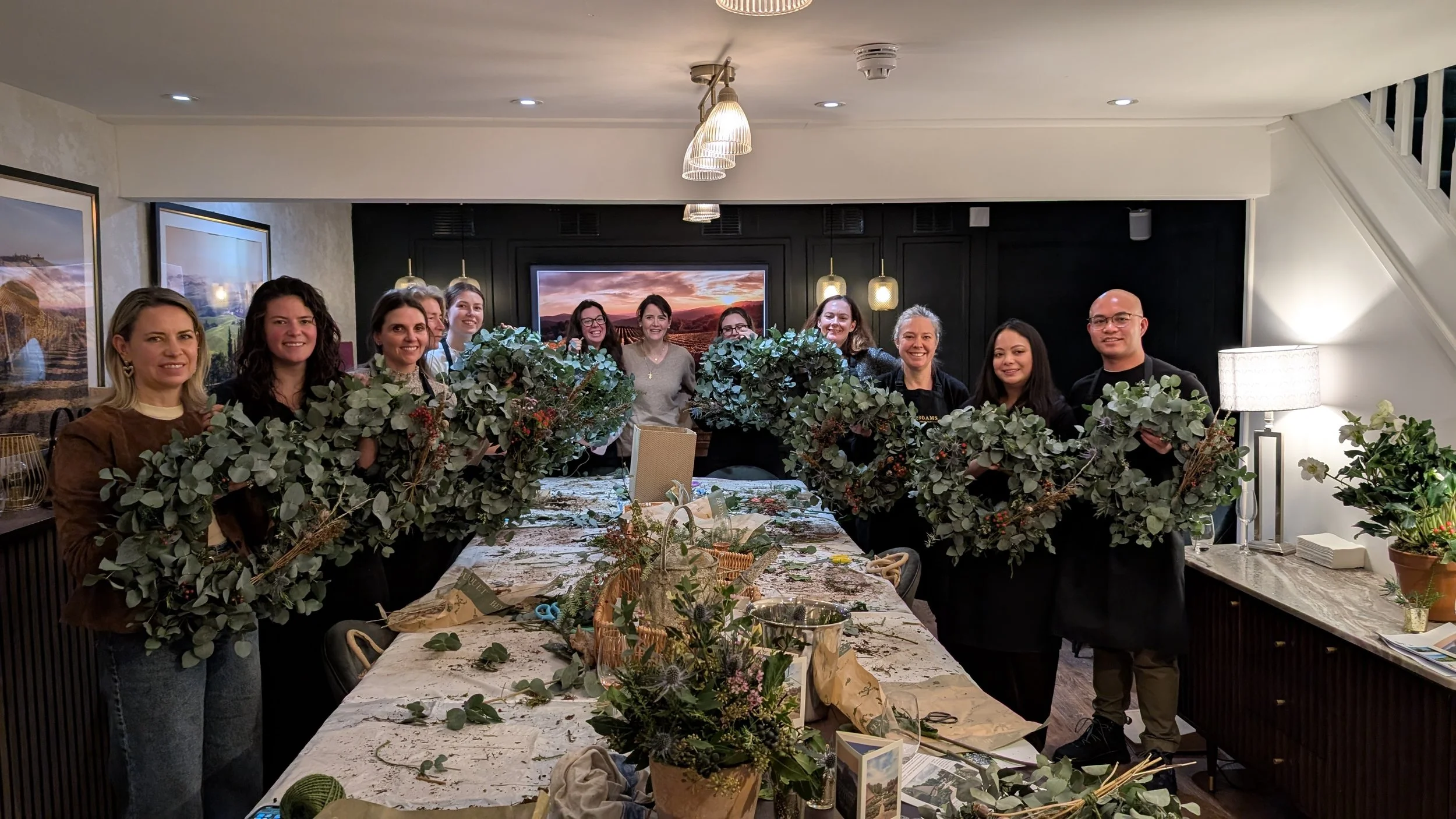 A group of people standing around a large table filled with green foliage and floral arrangements in a cozy, well-lit room, smiling at the camera.