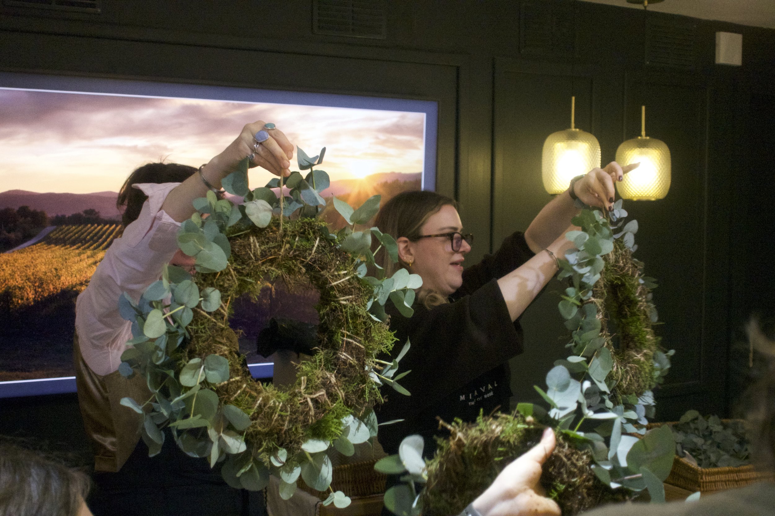 People gathered in a dark room, holding a large, circular arrangement of green eucalyptus leaves and moss.