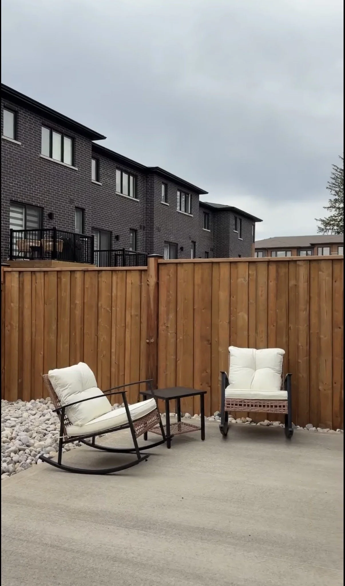 A backyard patio with two cushioned chairs, a small table, and a wooden privacy fence. The patio has a gravel area and a concrete section, with gray residential buildings in the background and an overcast sky.