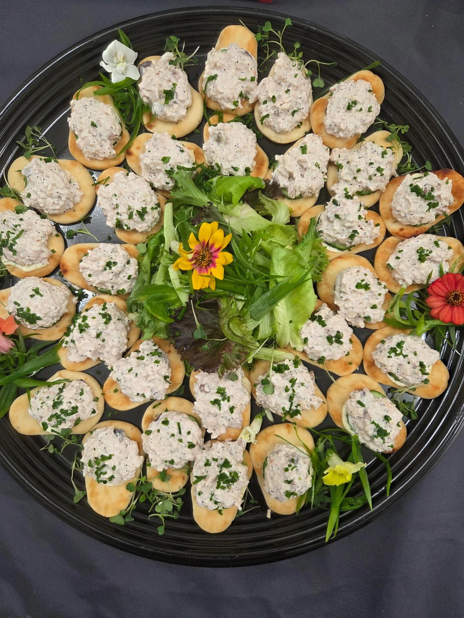 Tray of individual crackers topped with a creamy tuna salad garnished with chopped chives, surrounded by mixed greens and decorative flowers.
