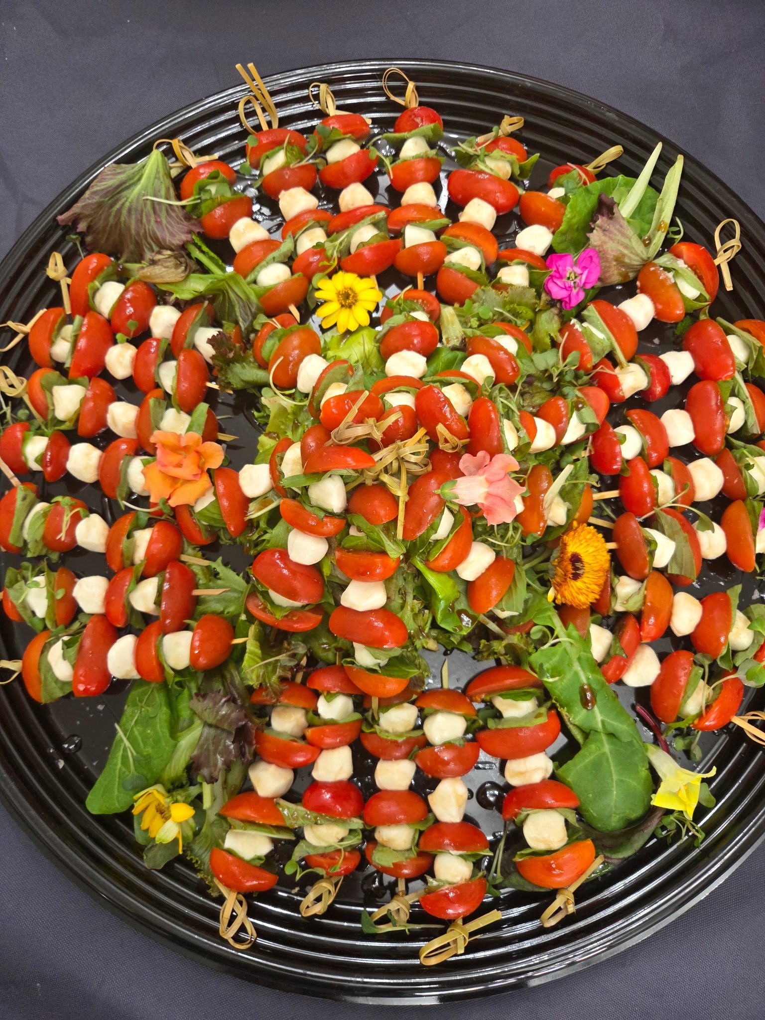 Plate with skewered appetizers featuring cherry tomatoes, mozzarella balls, basil, and edible flowers on a black platter.