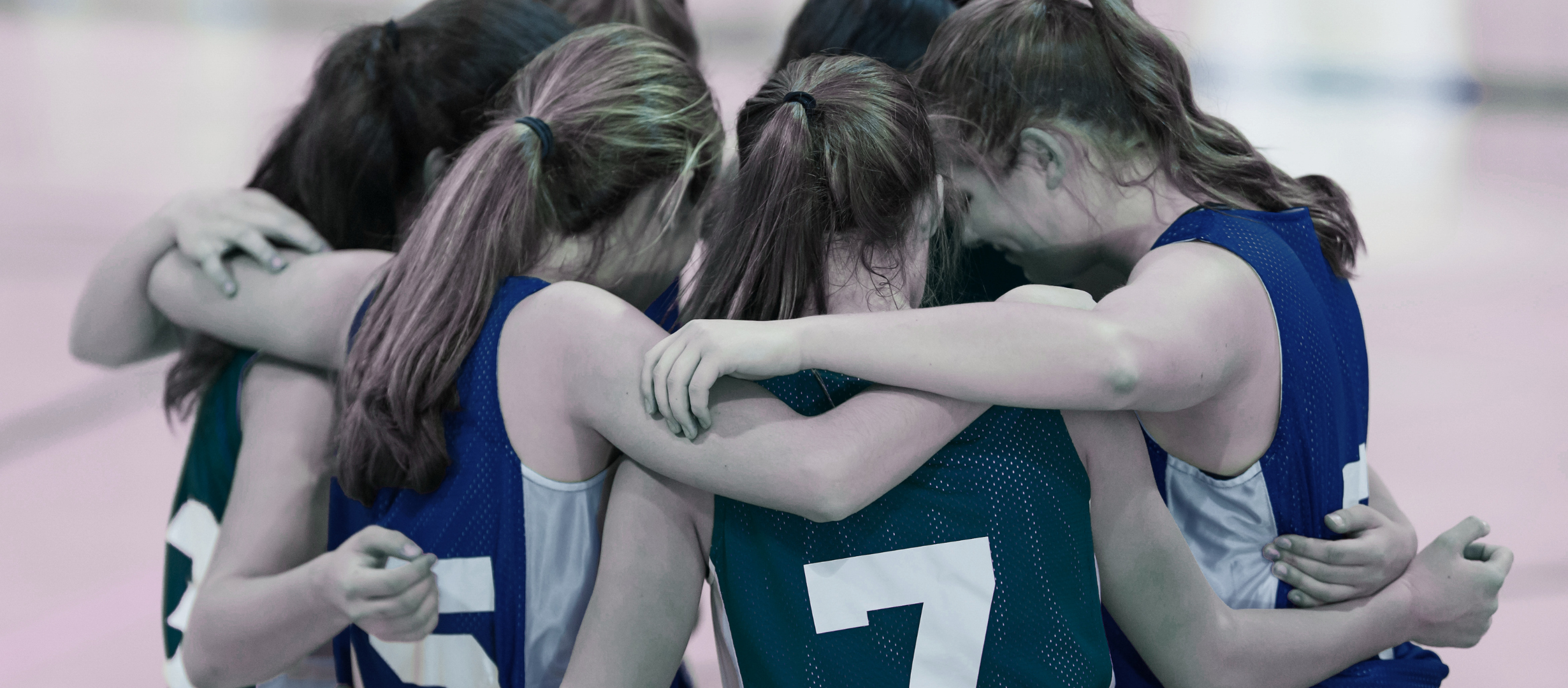 Group of female athletes in blue jerseys huddled together in a sports huddle, heads bowed and arms around each other.