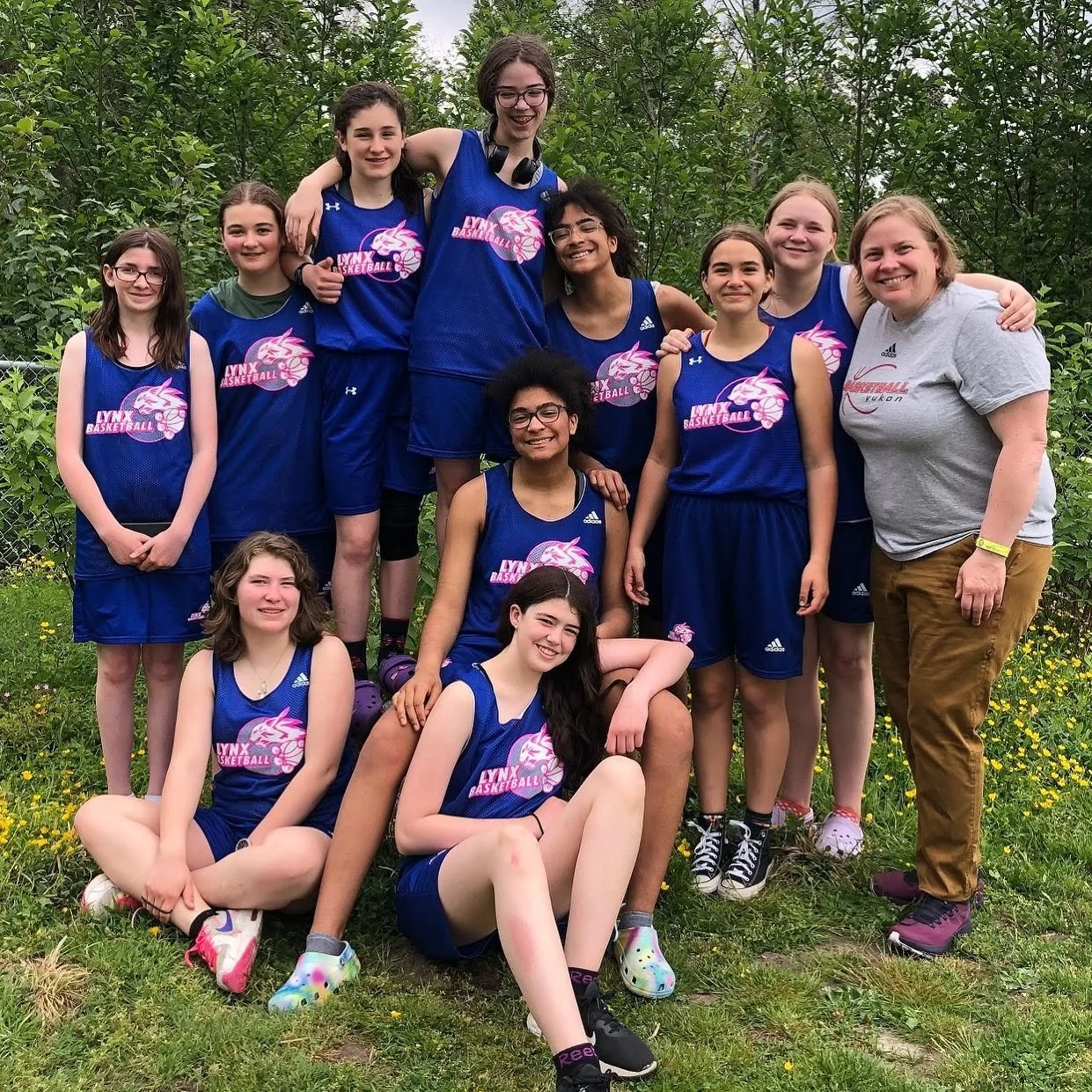 A girls' basketball team with their coach posing outdoors. The team members are wearing matching blue uniforms. They are standing and sitting on a grassy area with trees in the background, smiling at the camera.