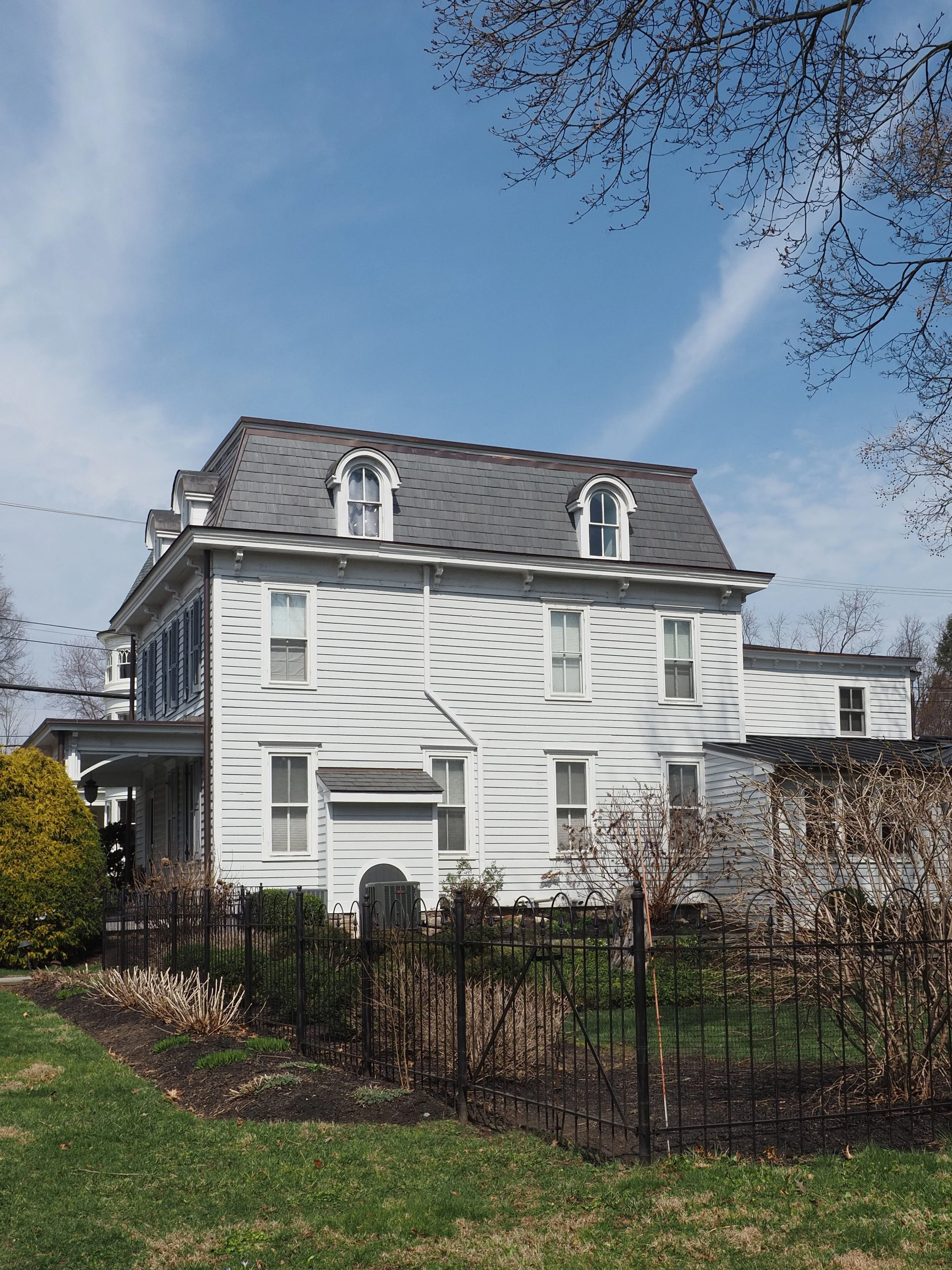 A large white Victorian-style house with three dormer windows on the roof, surrounded by a small garden with some bushes and trees, under a partly cloudy blue sky.