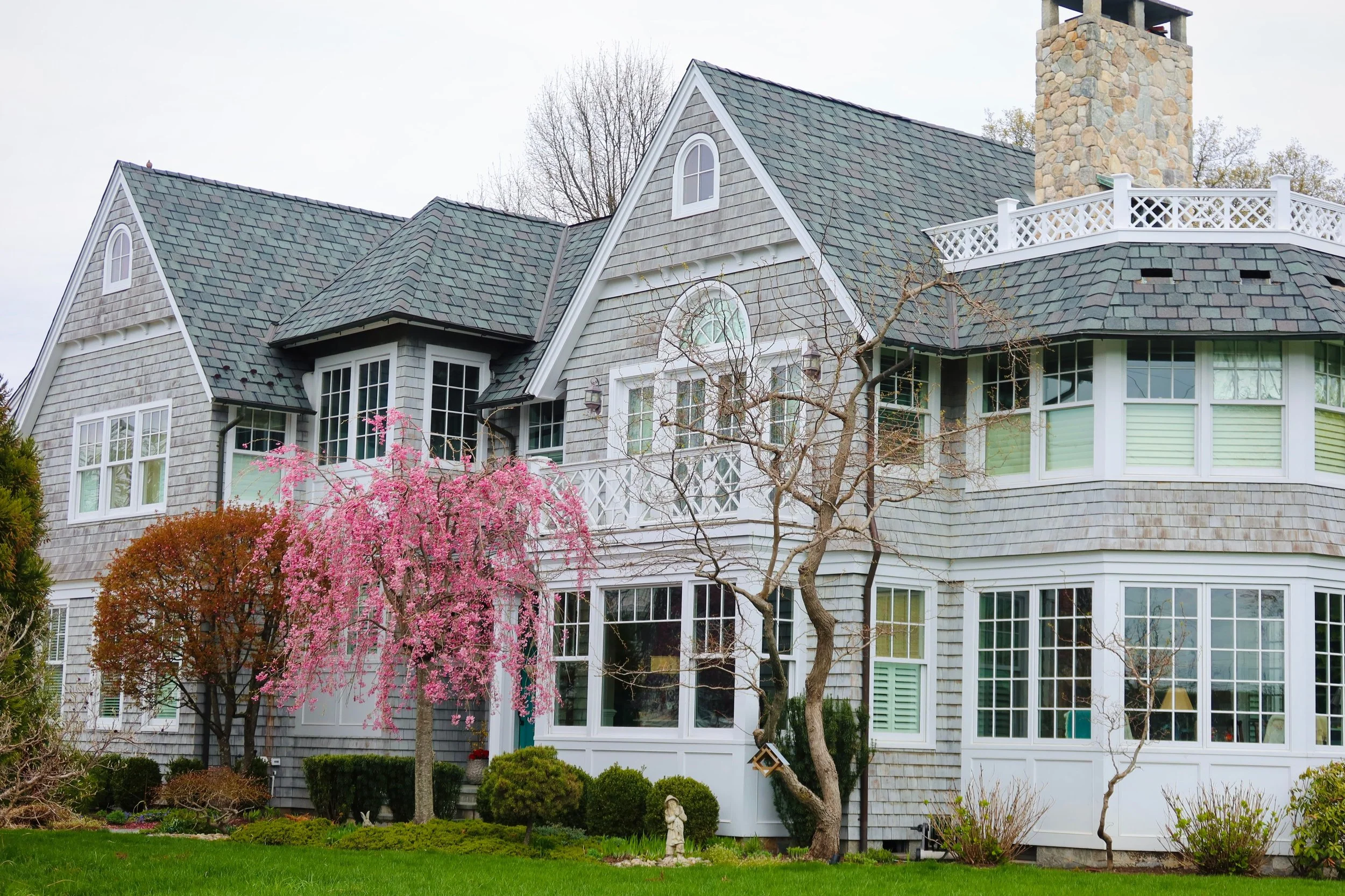 A large house with gray shingles, white trim, multiple gabled roofs, and a stone chimney. There are trees with pink blossoms, green grass, and a garden in front.